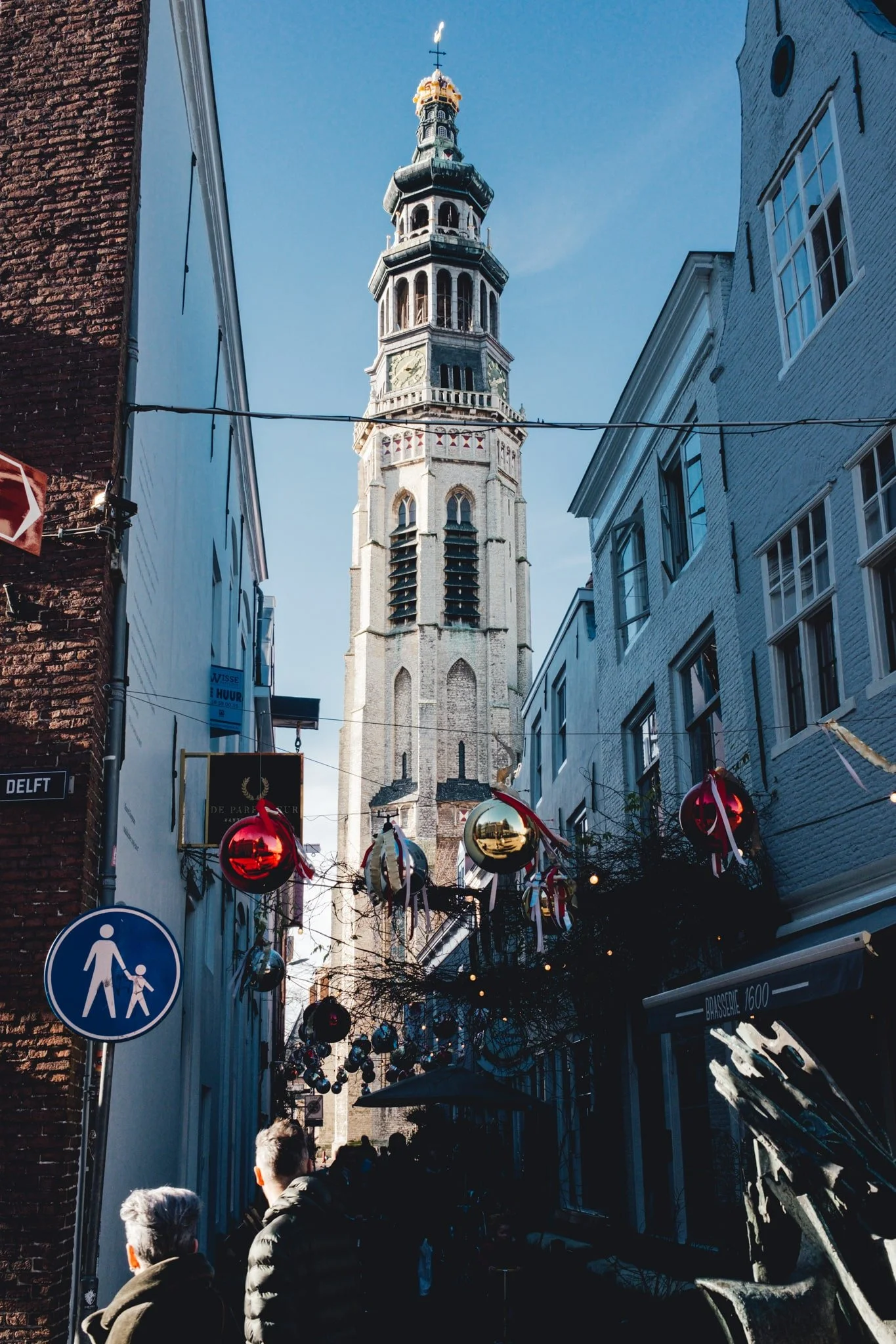 Uitzicht op een historische kerktoren in een straat met winkel- en horecagelegenheden, versierd met kerstballen en linten, onder een heldere blauwe hemel.