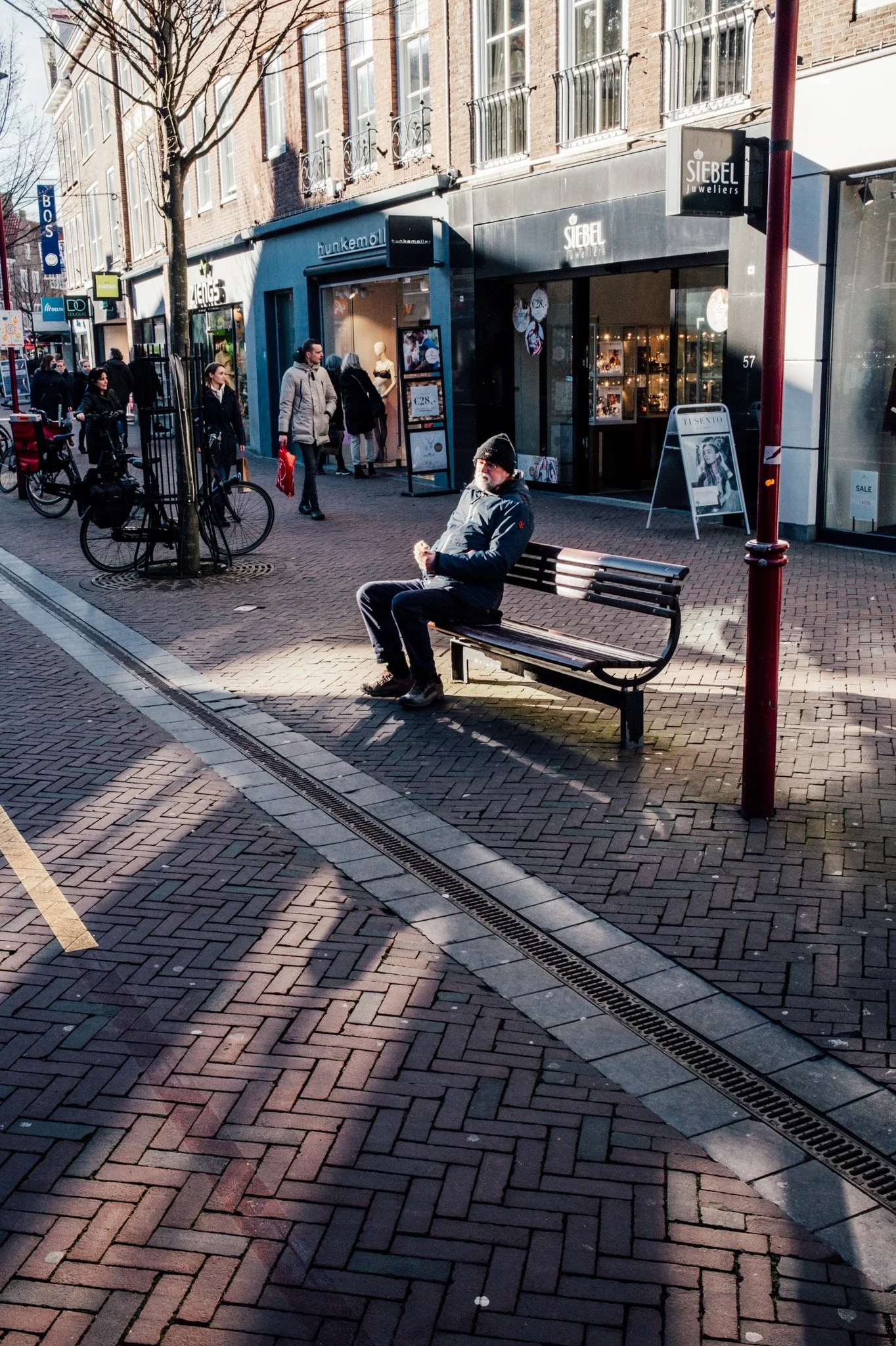 Man zit op een bankje op een stadsplein, met winkels op de achtergrond zoals juweliers en boetieks, onder een boom en in de zon.