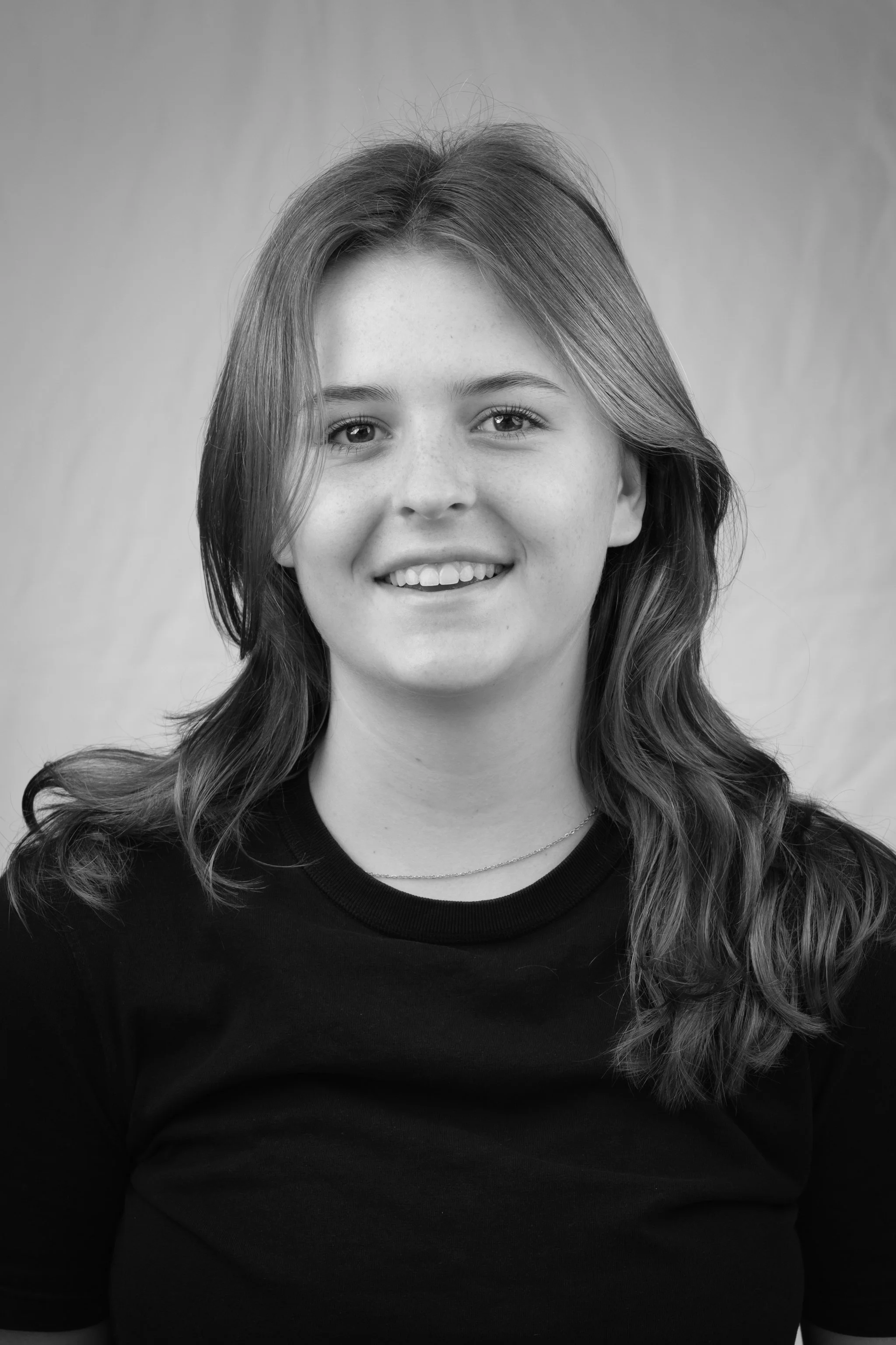 Zwart-wit portret van een jonge vrouw met lang, lichtbruin haar, lachend, gekleed in een zwarte t-shirt, tegen een effen achtergrond.