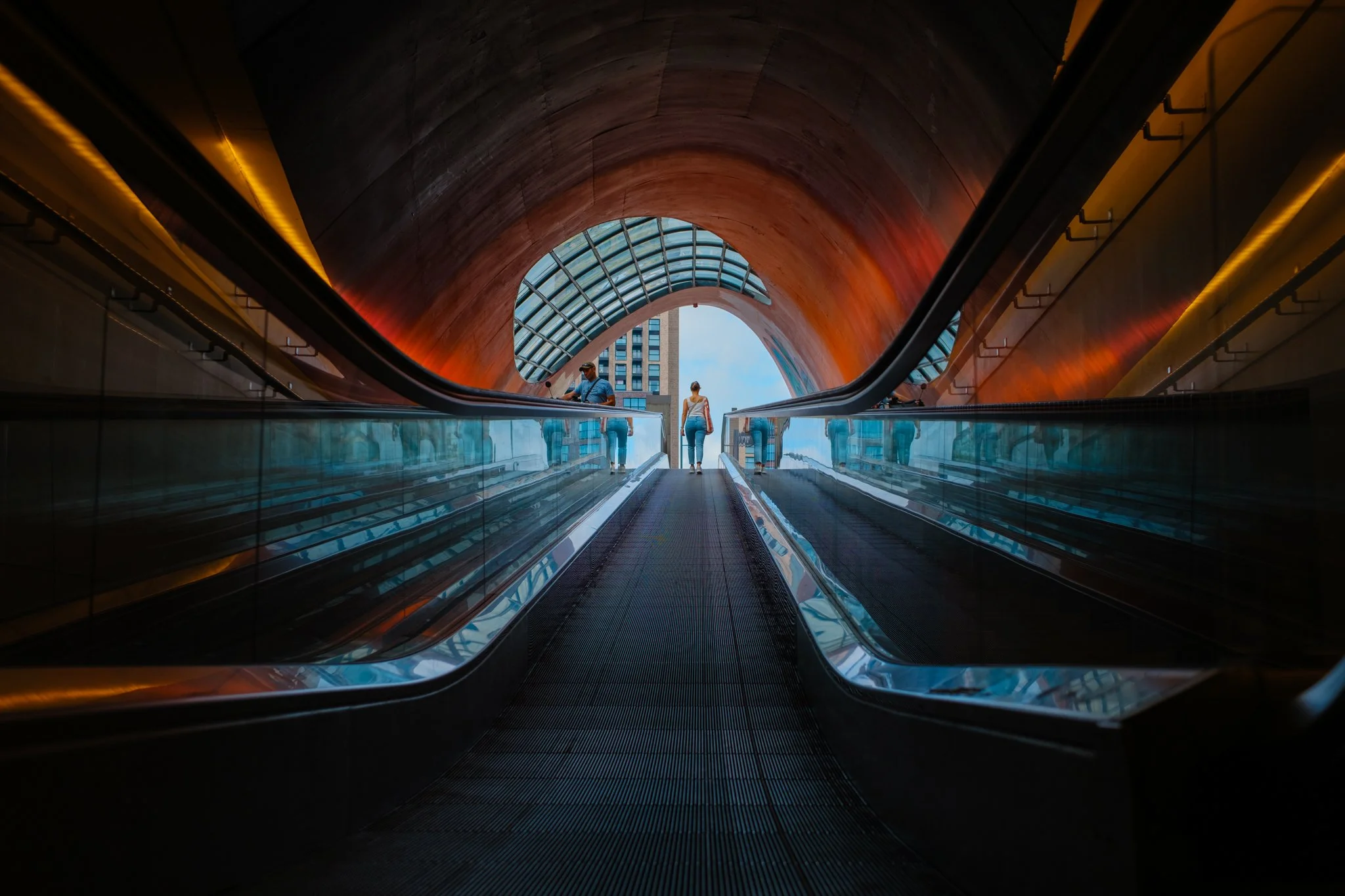 Twee mensen op een roltrap in een modern, architectonisch gebouw met een ronde, houten binnenboog en glazen muur.