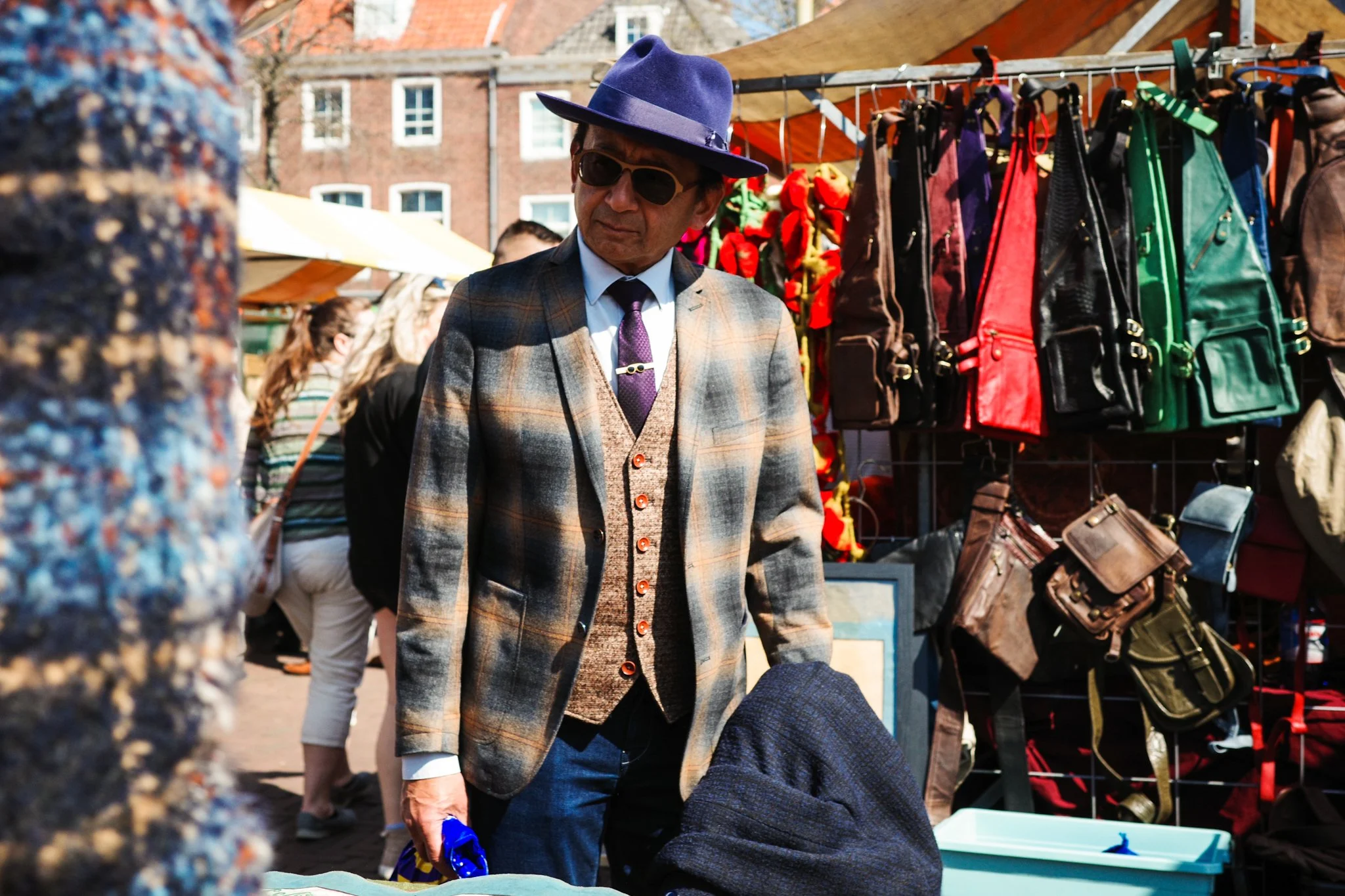 Man in een geruit jasje en hoed op een markt, met tassen en kleding op de achtergrond.