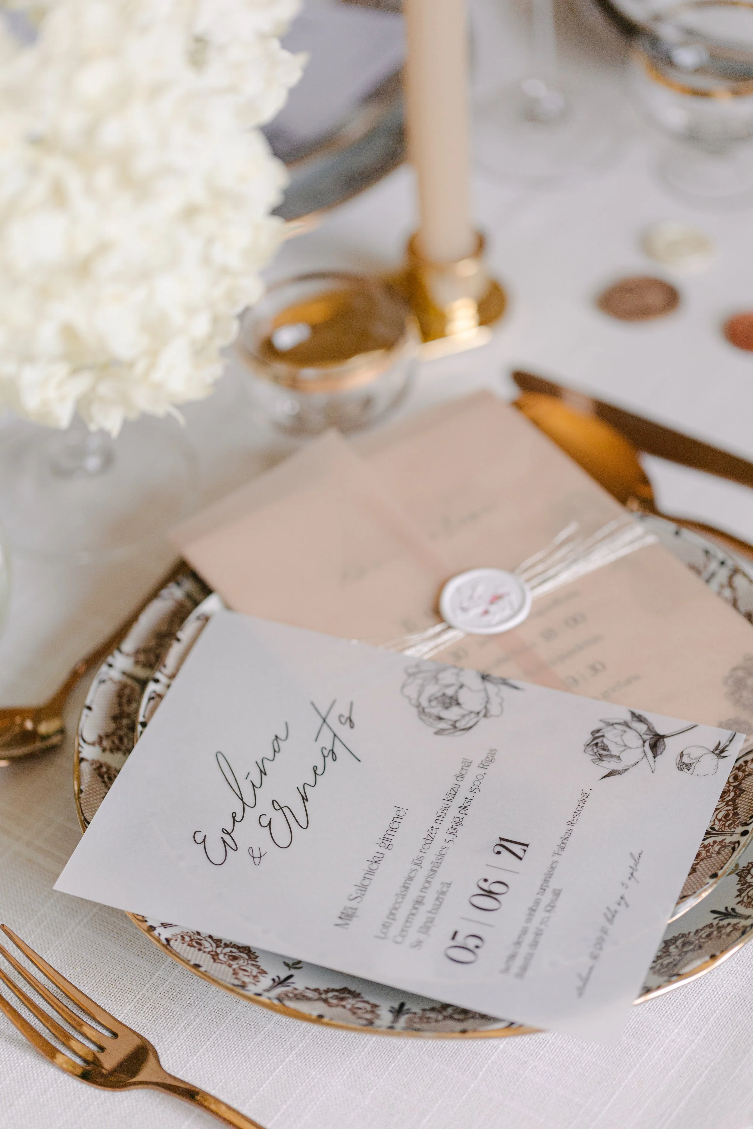 Wedding invitation card on decorative plate, set on a table with gold utensils, white floral centerpiece, and candle.