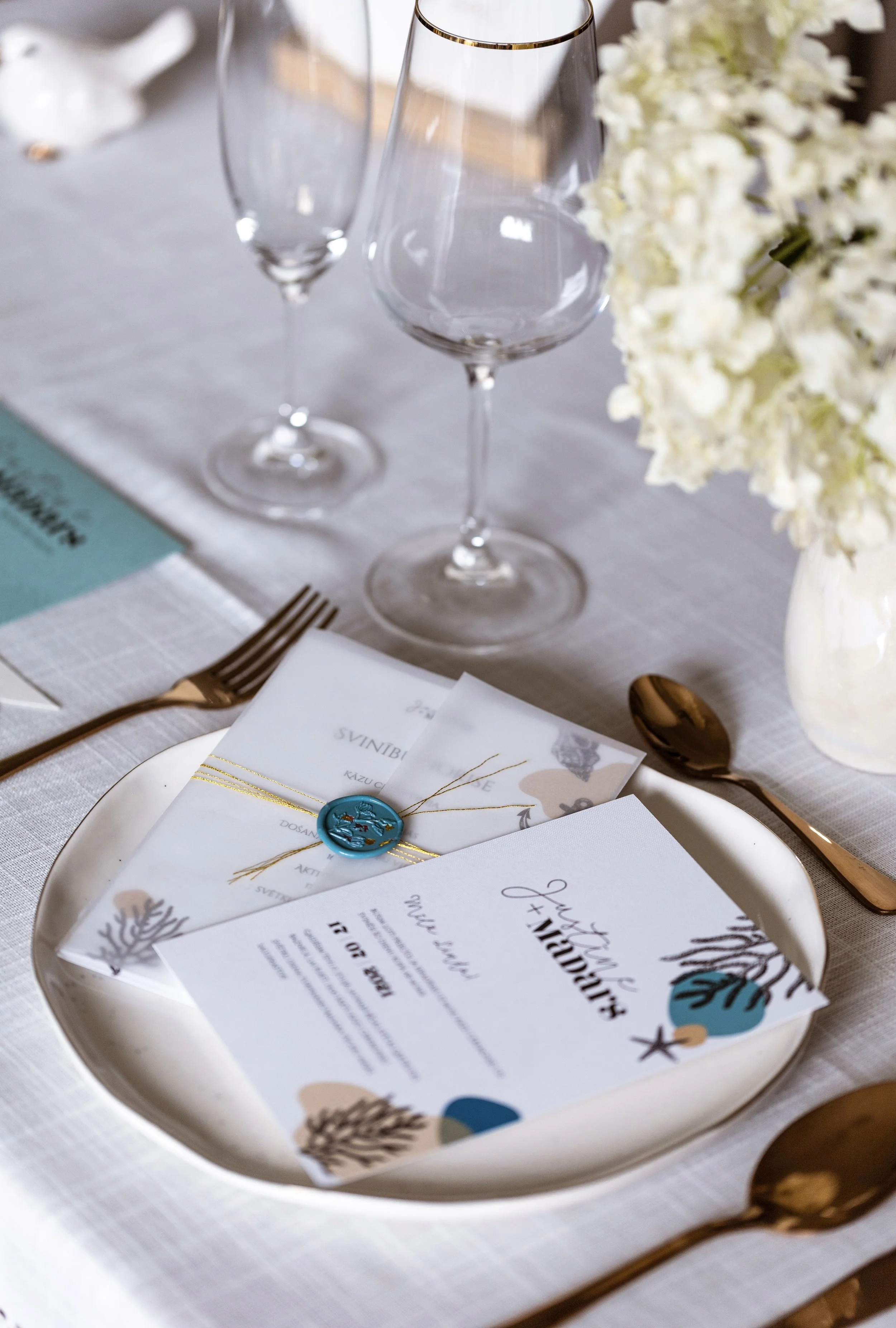Elegant table setting with white plates, gold utensils, wine glasses, floral napkins with a tied blue wax seal, and a menu with a beach-themed design, on a white tablecloth.