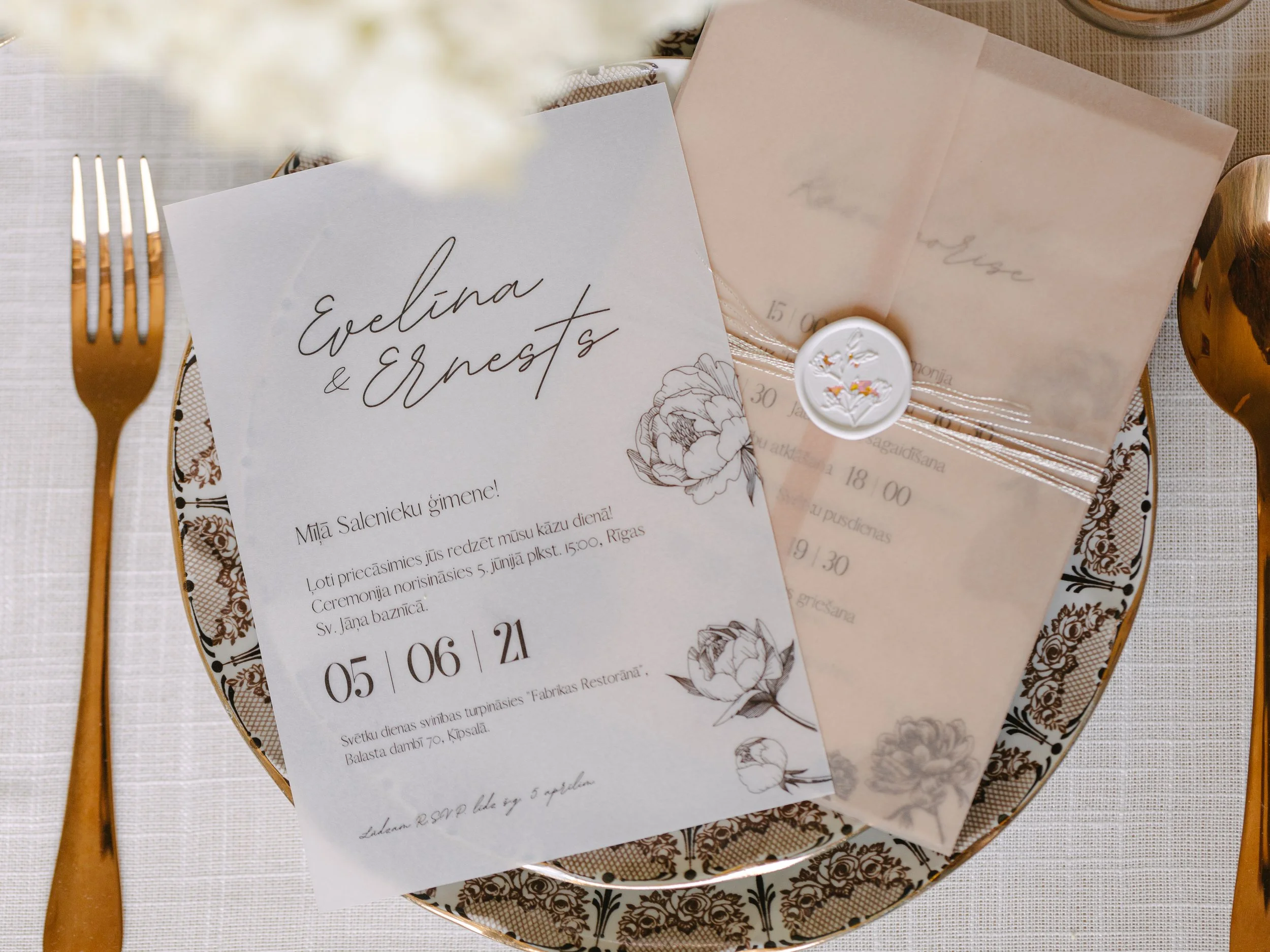 Invitation card for Evelina and Ernests wedding placed on a decorative plate. The card features floral illustrations and details about the wedding date and location. Gold-colored cutlery is positioned on a white tablecloth.