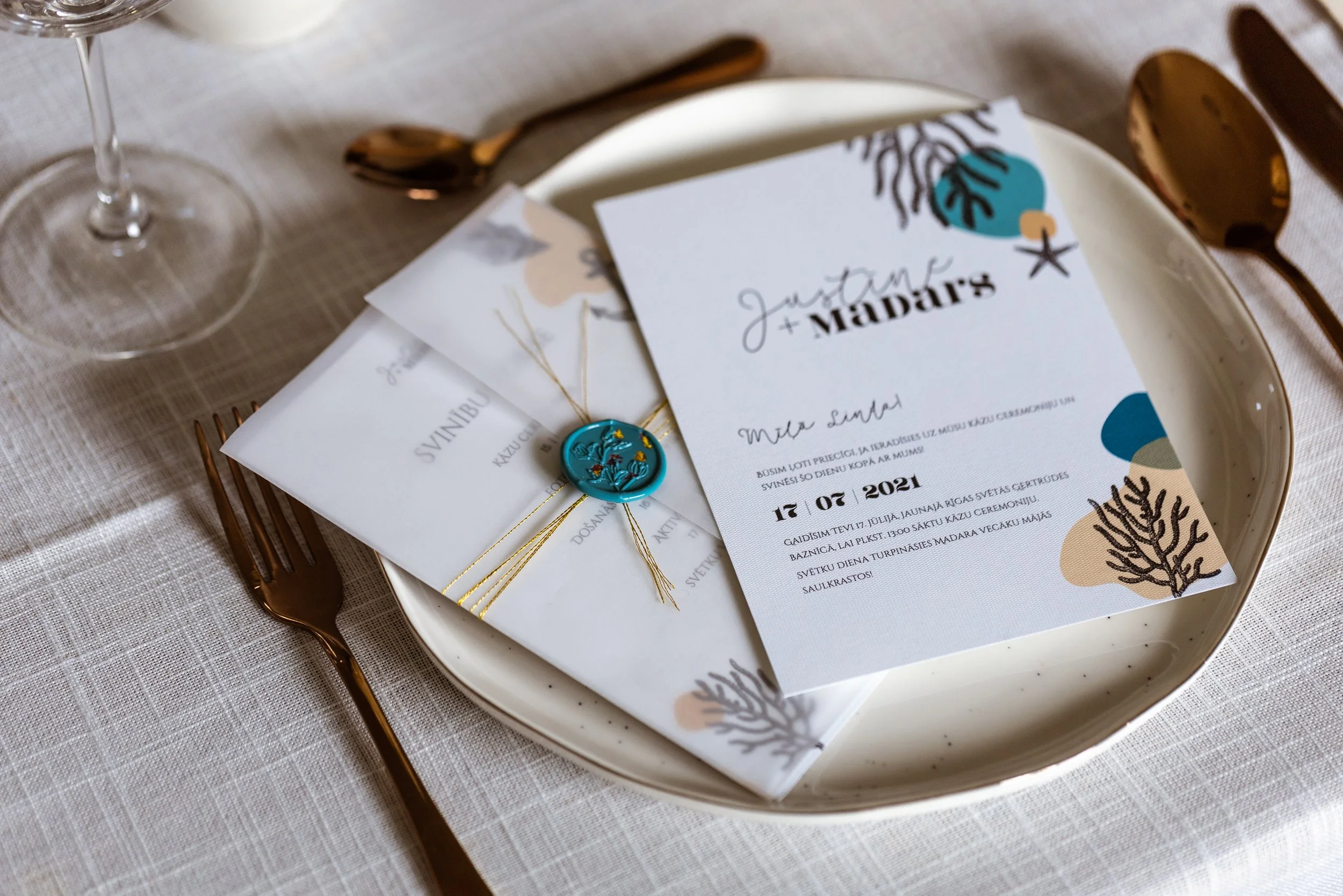 Wedding invitation and envelope on a white plate with gold-colored fork and spoon, a wine glass, placed on a textured beige tablecloth.