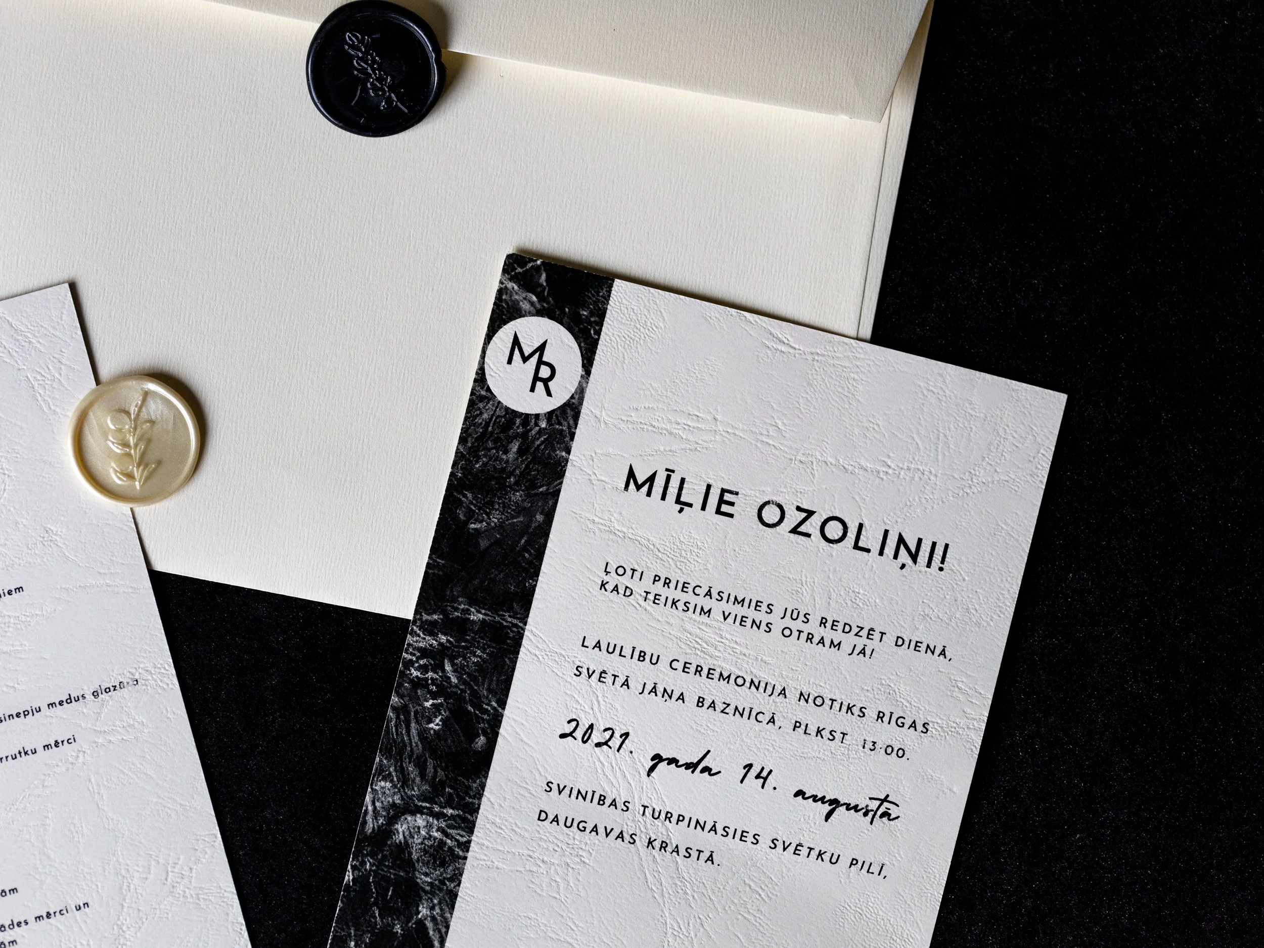 A wedding invitation card in Latvian language with black and white marble design and a handwritten font. It is placed on a dark surface with wax seals and additional stationery around it.