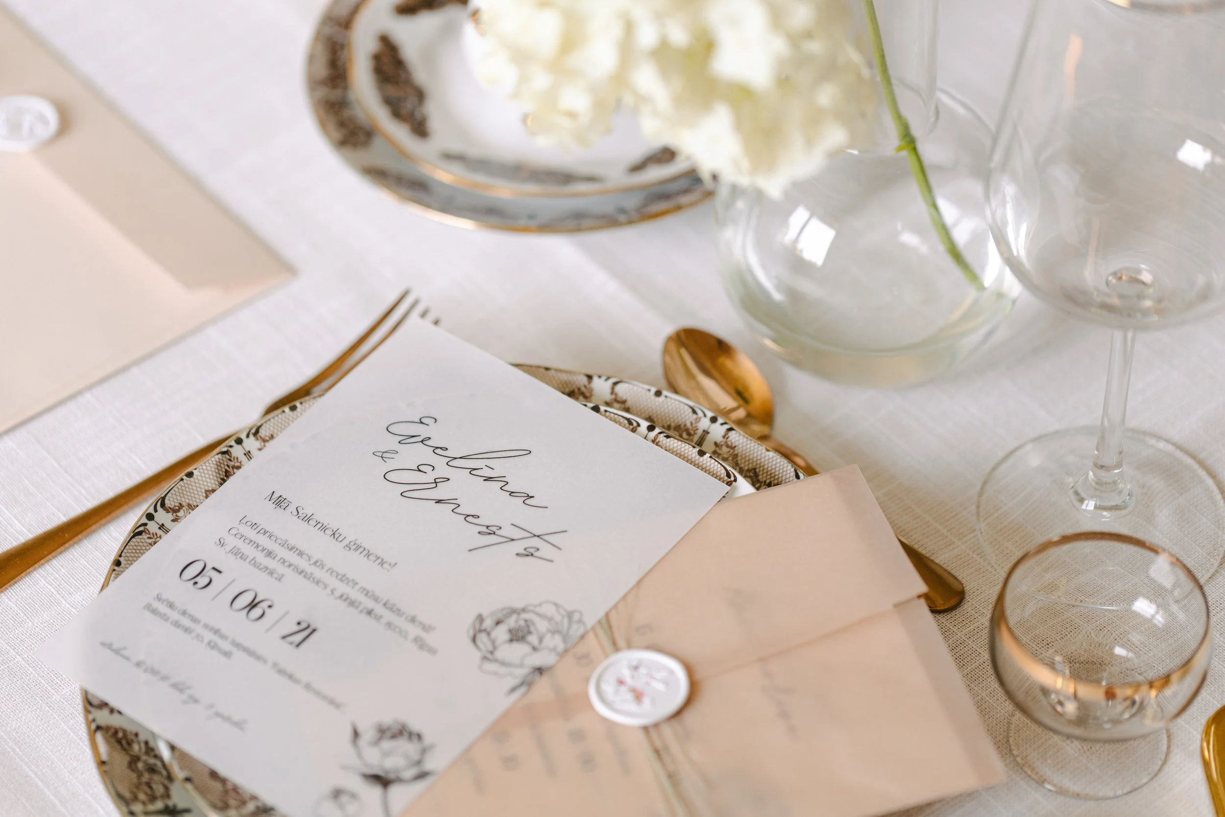 Wedding table setting with a menu card, gold cutlery, floral decoration, and empty glassware on a white tablecloth.