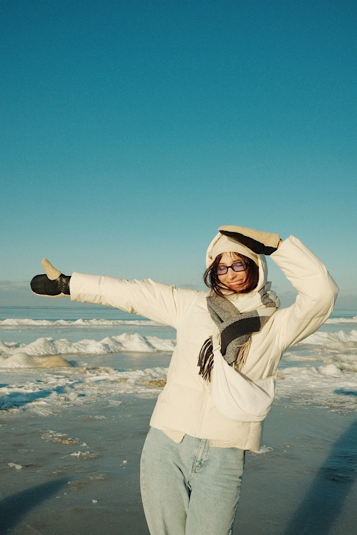 Woman in winter clothing, sunglasses, and gloves on a snowy beach, smiling with arms outstretched.
