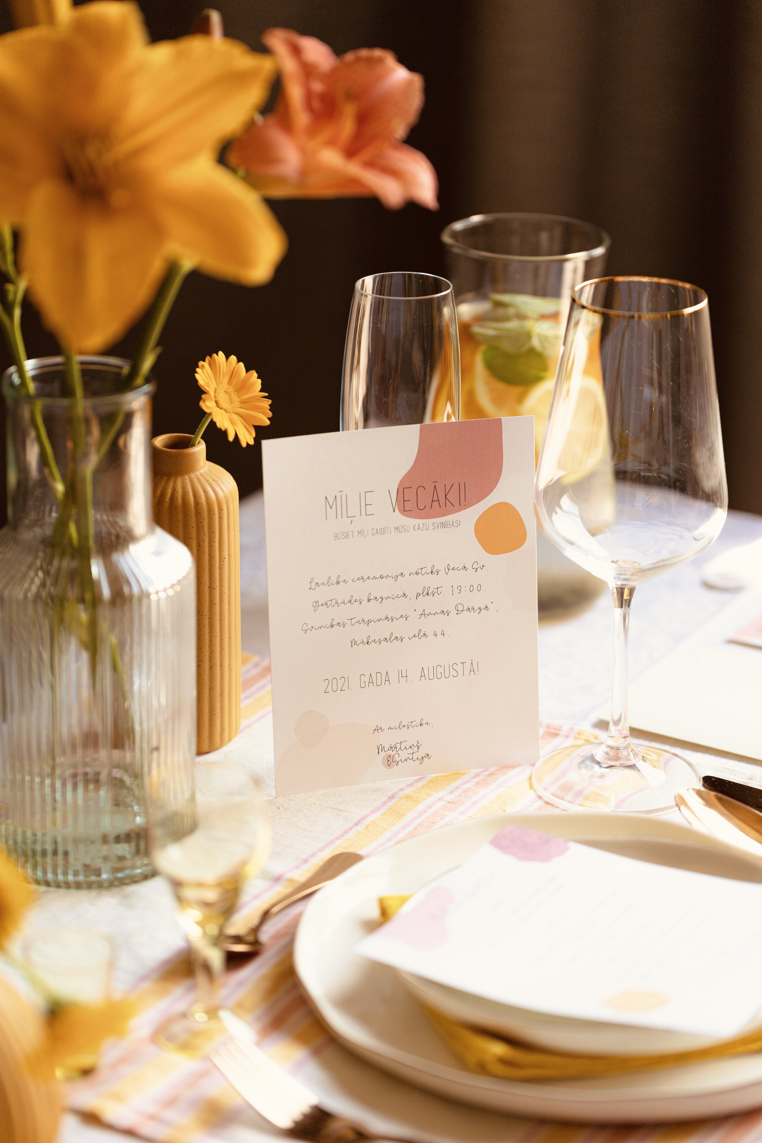 A table setting with a bouquet of flowers, two empty wine glasses, a pitcher with citrus fruit slices and mint, a plate with a menu, and a card for a celebration on a striped tablecloth.