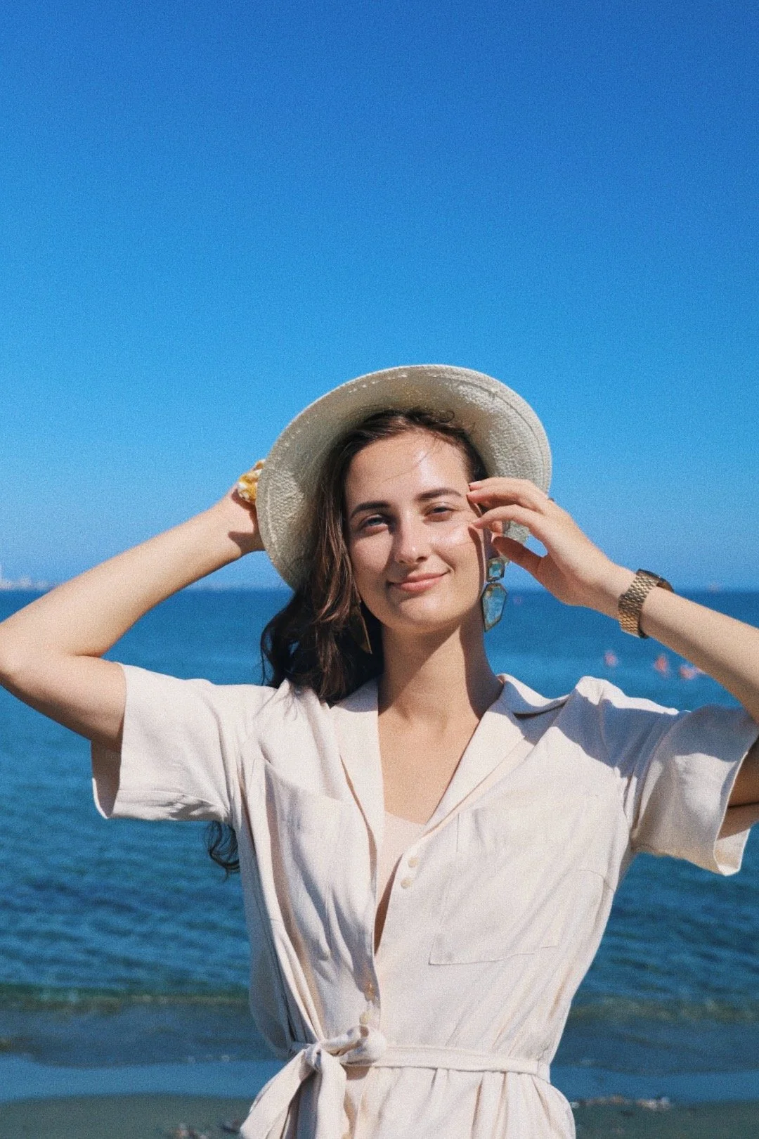 Woman in a white dress and straw hat standing on a beach with ocean and clear blue sky in the background.