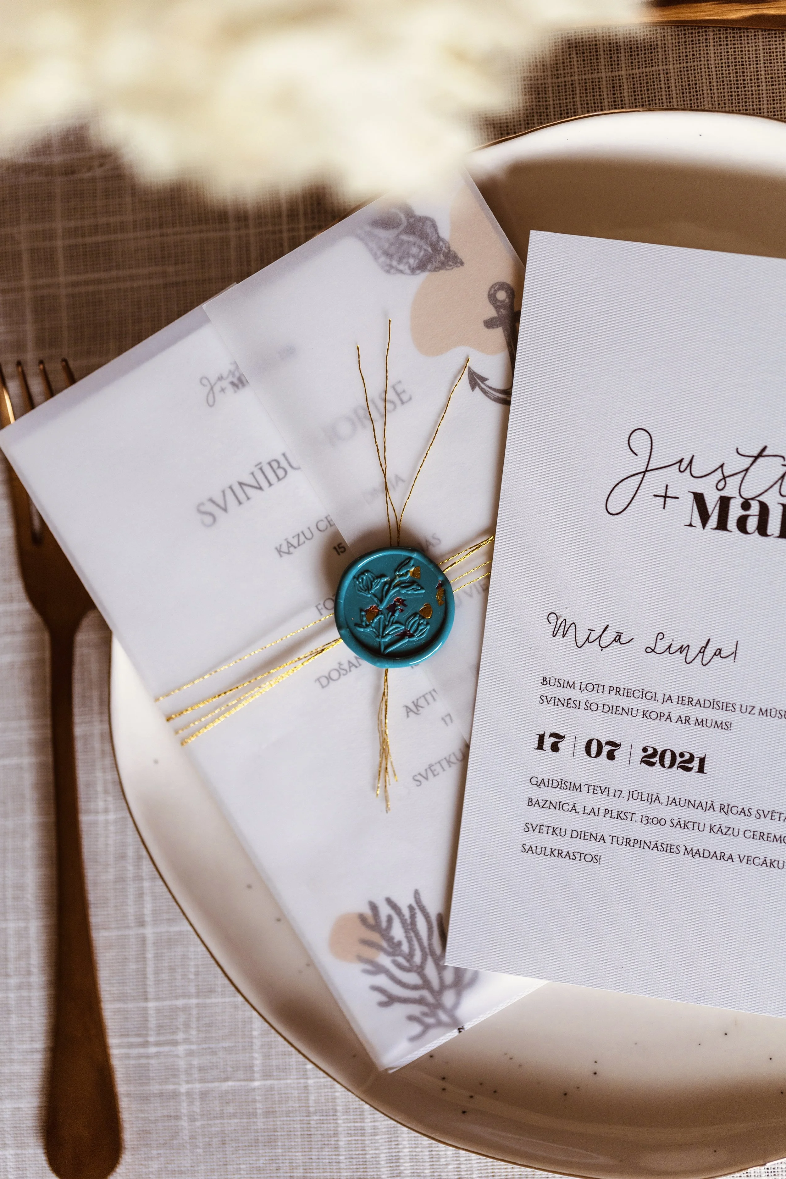 Invitation for a wedding celebration with a close-up of a printed card and a wrapped item tied with gold string and a blue wax seal, placed on a white plate.