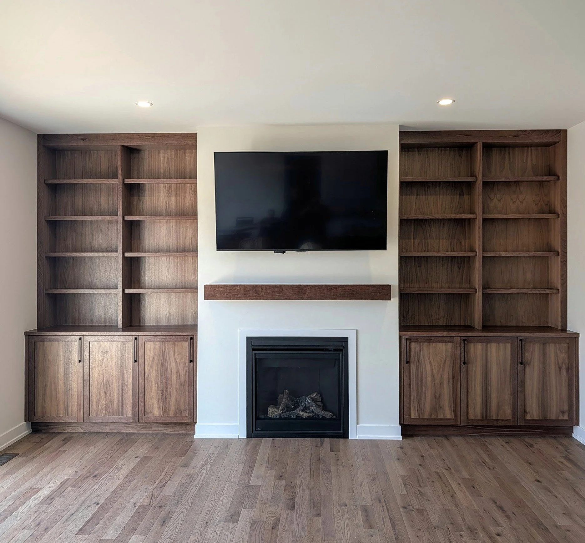Living room with built-in wooden bookshelves on either side of a TV mounted above a fireplace with a wooden mantel on a white wall, and a hardwood floor.