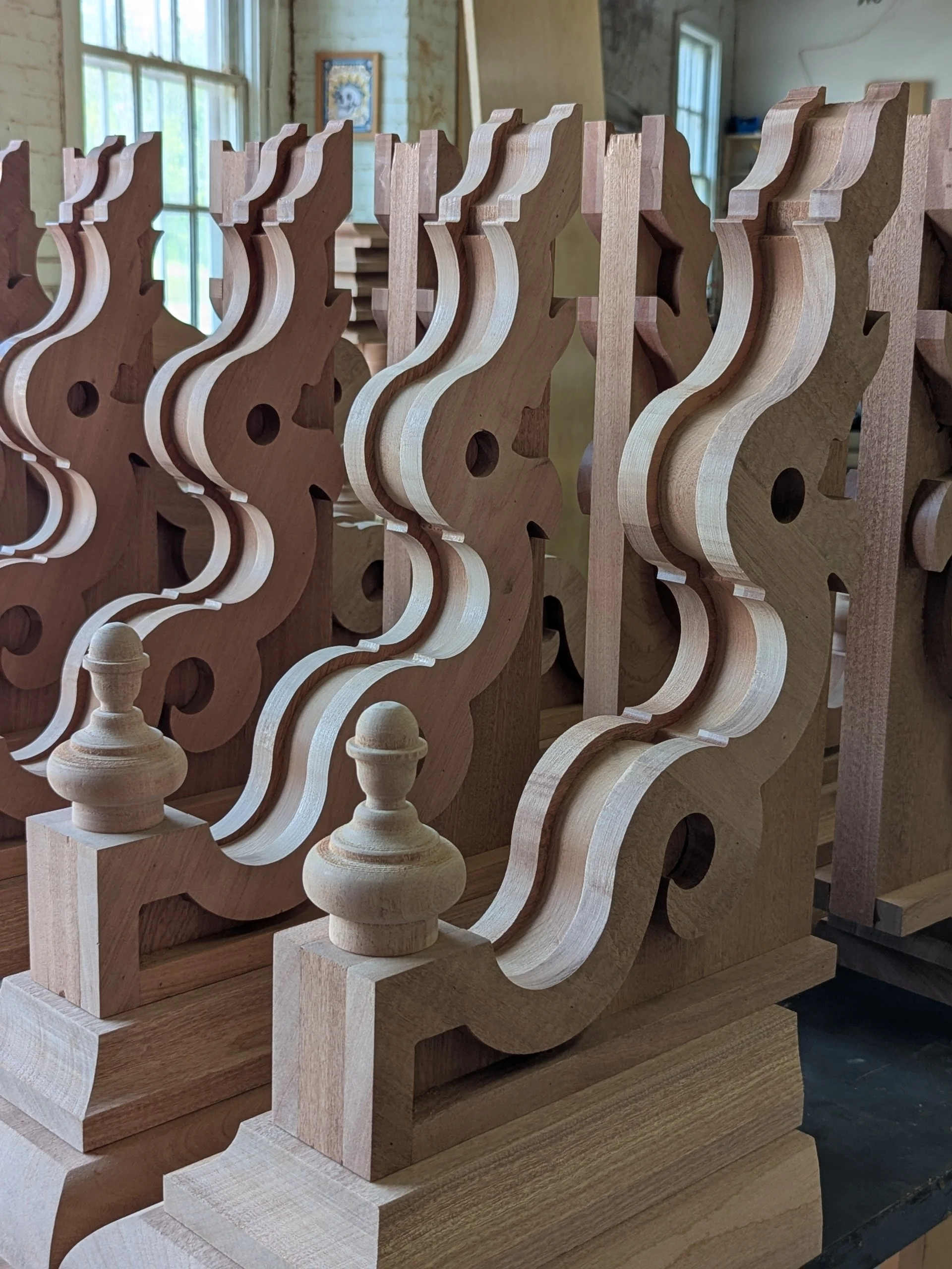 Wooden chess piece stands in front of carved wooden chessboard frames in a woodworking shop with large windows, a framed picture on the wall, and natural light.