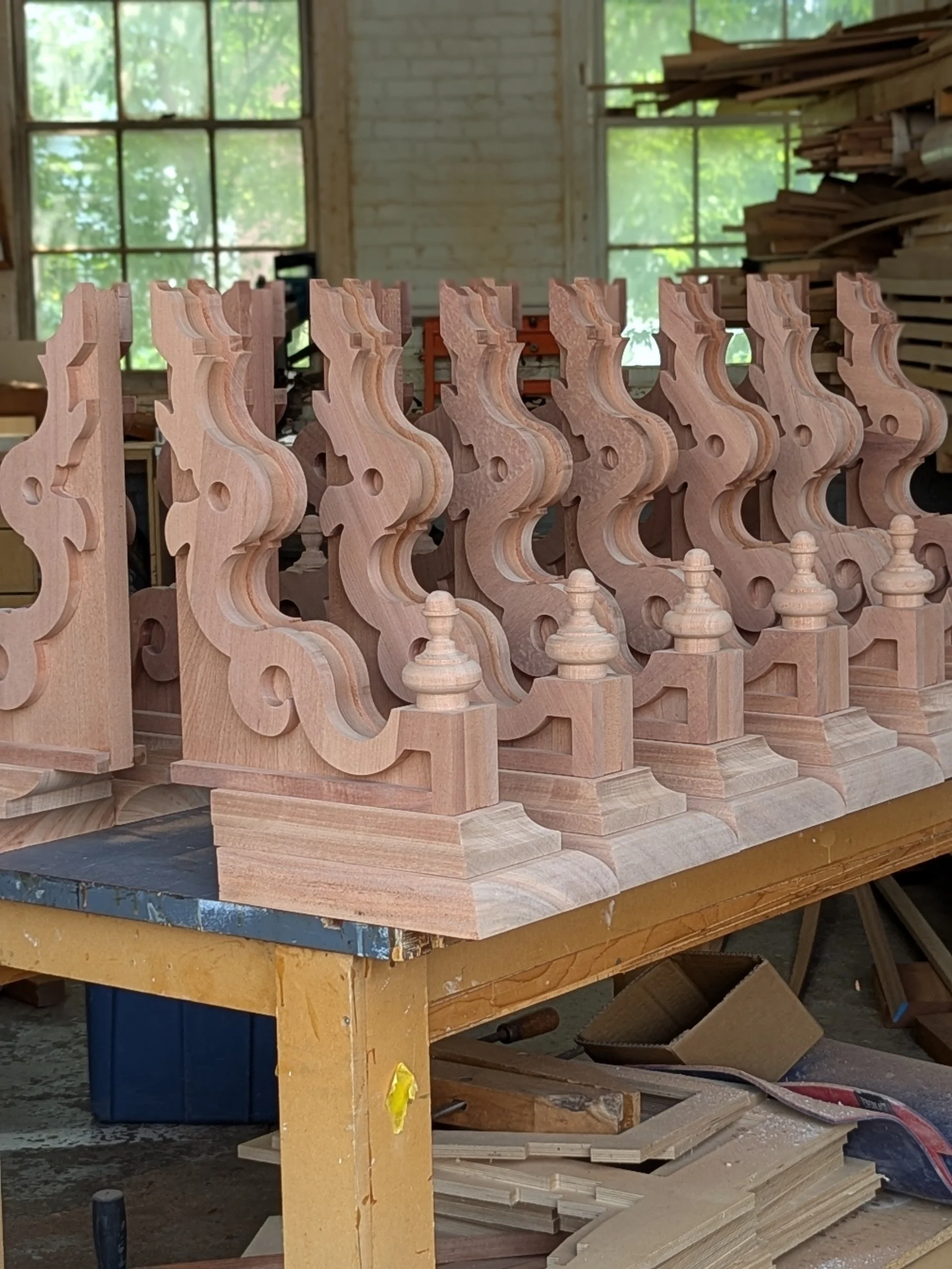 Several unfinished wooden decorative posts arranged on a workbench inside a woodworking shop with large windows and stacks of wood in the background.