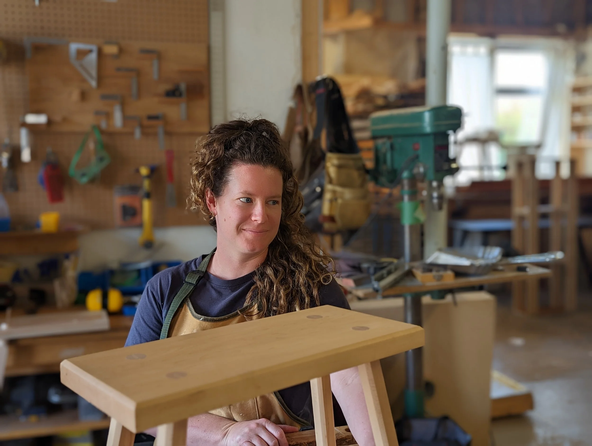 A woman in a woodworking shop looking at a small piece of wooden furniture she is working on.