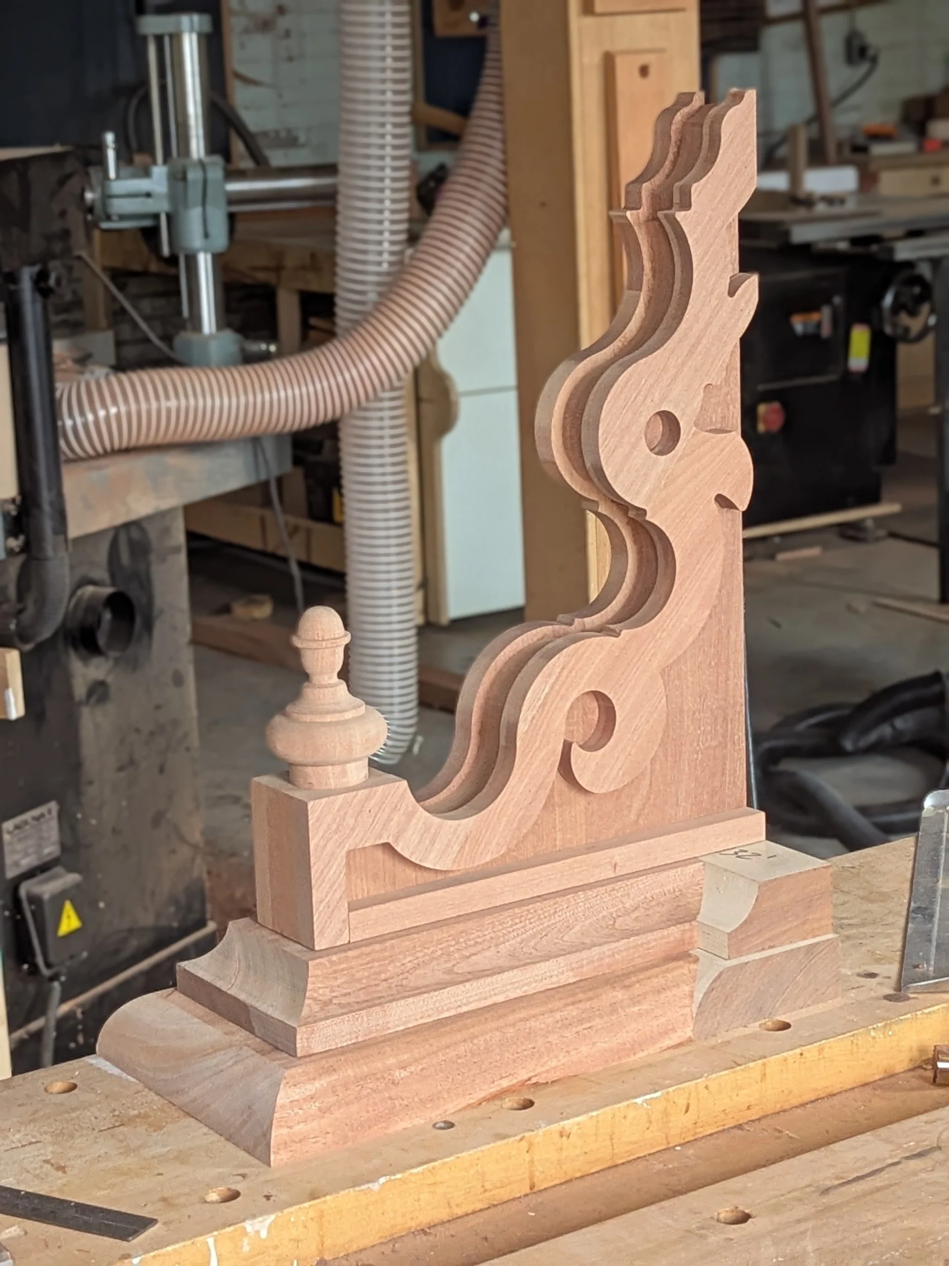 A detailed wooden scrollwork piece in a woodworking shop, with various machinery and tools in the background.
