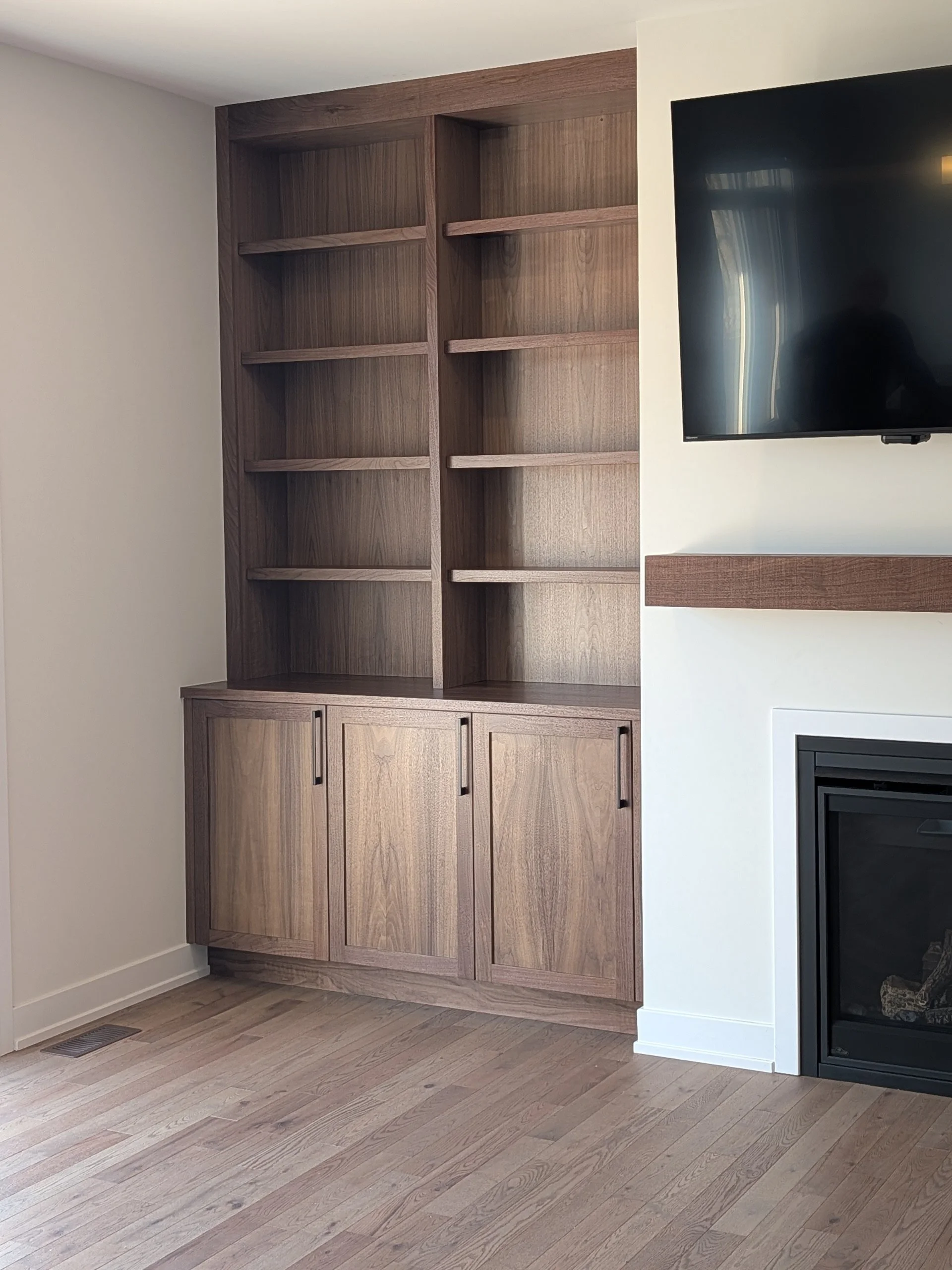 Empty wooden bookshelf with four shelves and cabinet doors at the bottom, adjacent to a mounted flat-screen TV and a fireplace, in a living room.