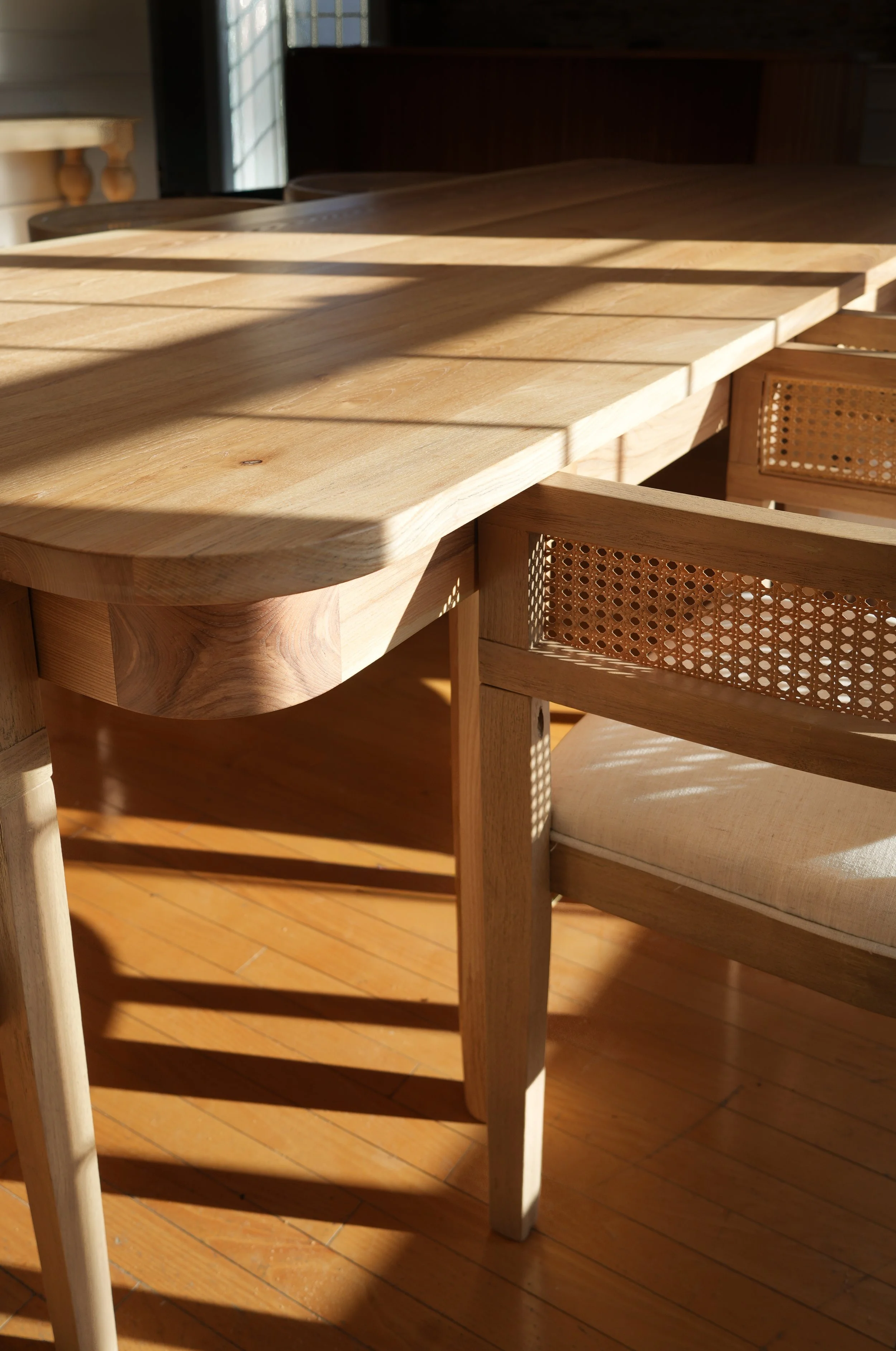 Close-up of a wooden dining table and chair with woven cane backrest, illuminated by sunlight through a window, casting shadows on a hardwood floor.