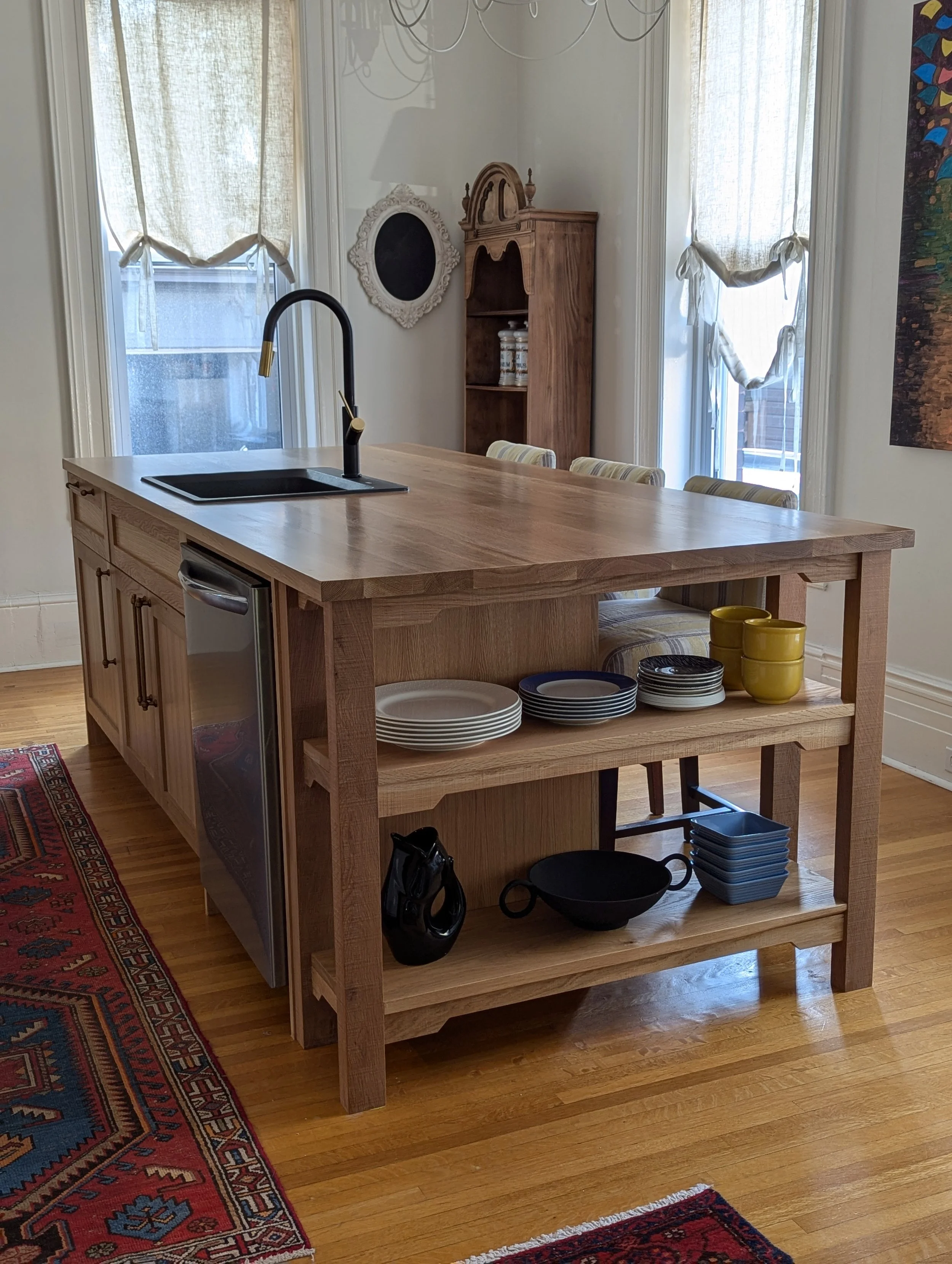 A kitchen island with a built-in sink and black faucet, displaying stacked plates and bowls, additional bowls and a jug on the shelf, with a window and a wooden cabinet in the background.