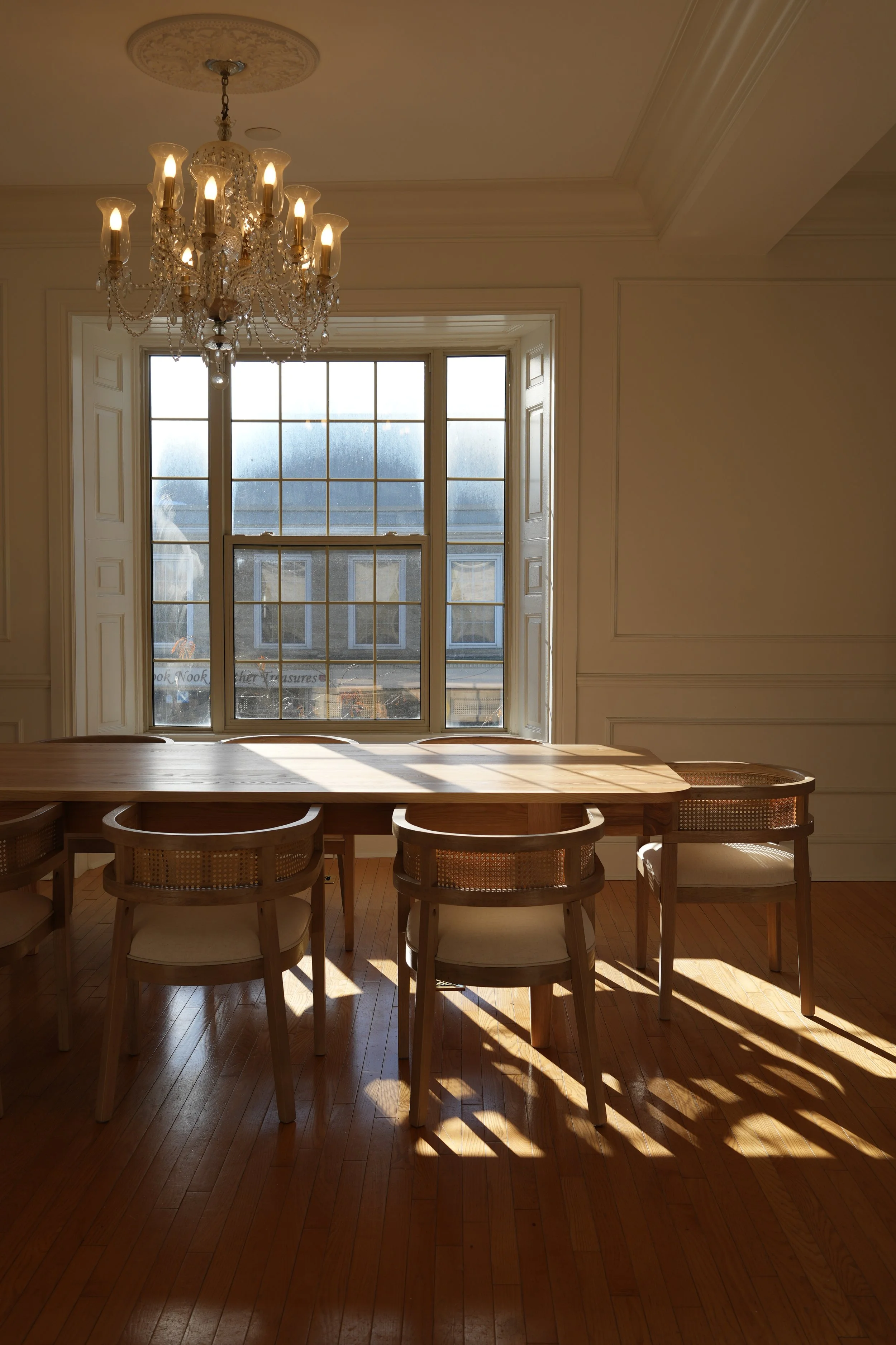 A dining room with a wooden table and six chairs, a large window with shutters, a chandelier hanging from the ceiling, and sunlight casting shadows on the hardwood floor.