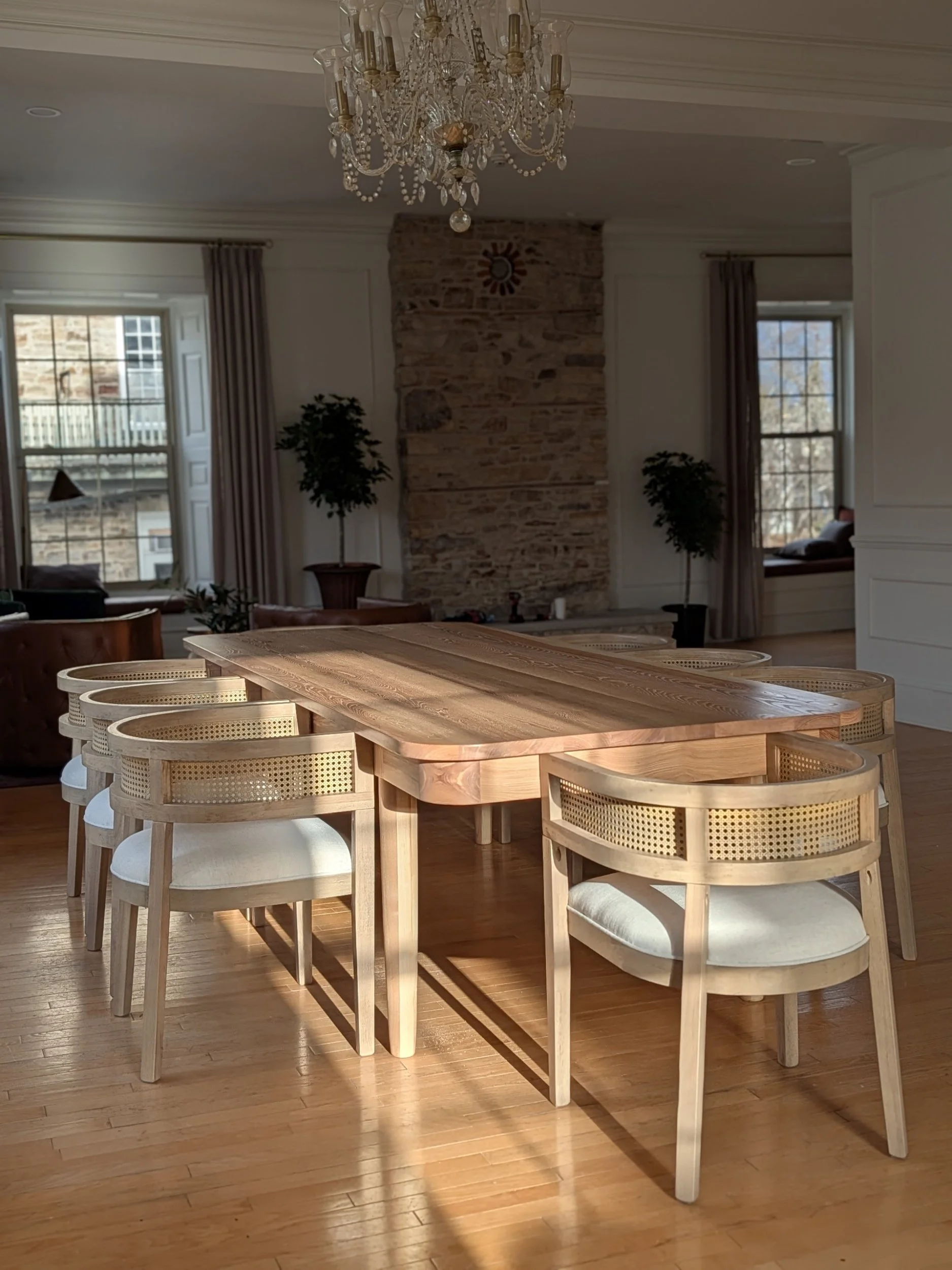 A wooden dining table with eight matching chairs in a spacious, well-lit room with hardwood floors, large windows, a stone fireplace, a chandelier, and potted plants.