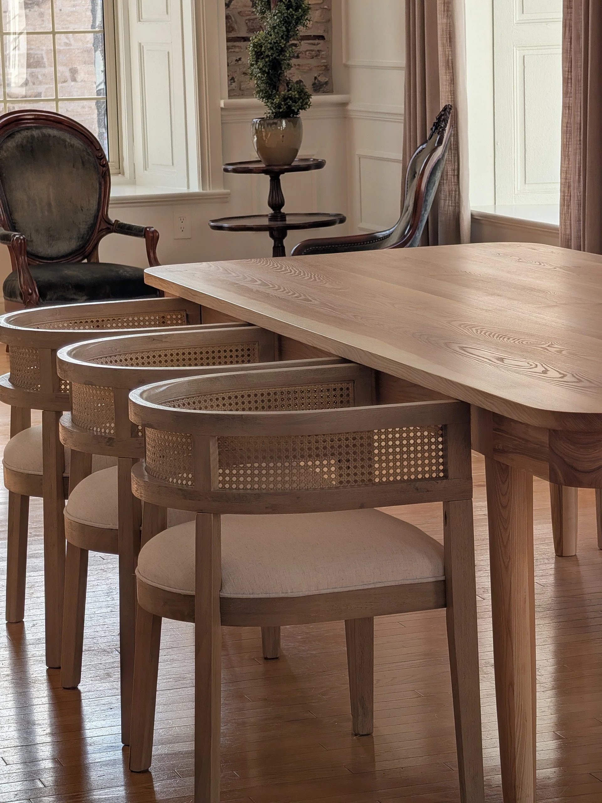 Wood dining table with four wooden chairs featuring woven cane backs and upholstered seats in a well-lit room with a stone wall, windows, curtains, and decorative plants.