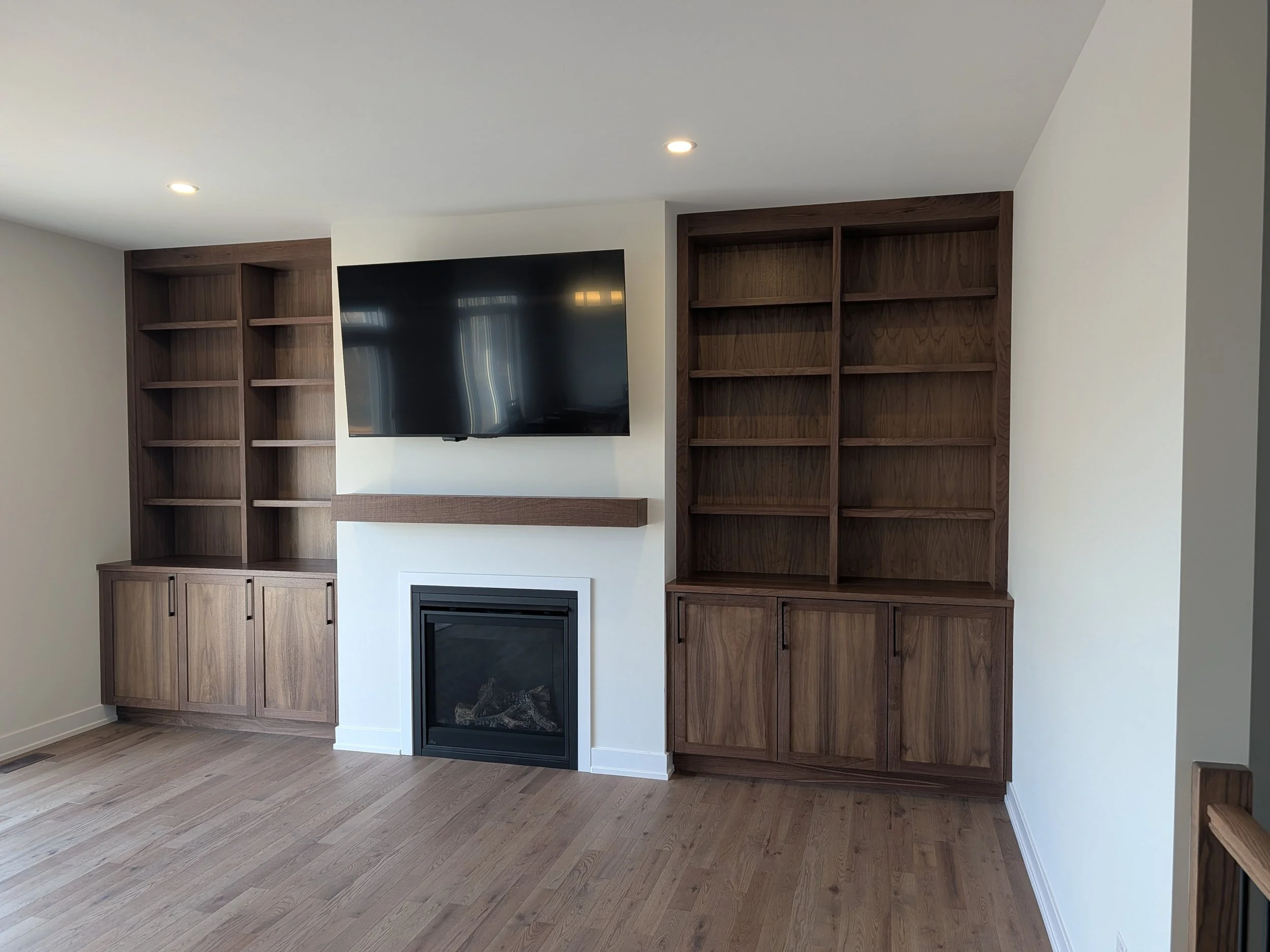 Living room with built-in wooden bookshelves, a mounted flat-screen TV, and a fireplace beneath the TV, with hardwood flooring and recessed ceiling lights.