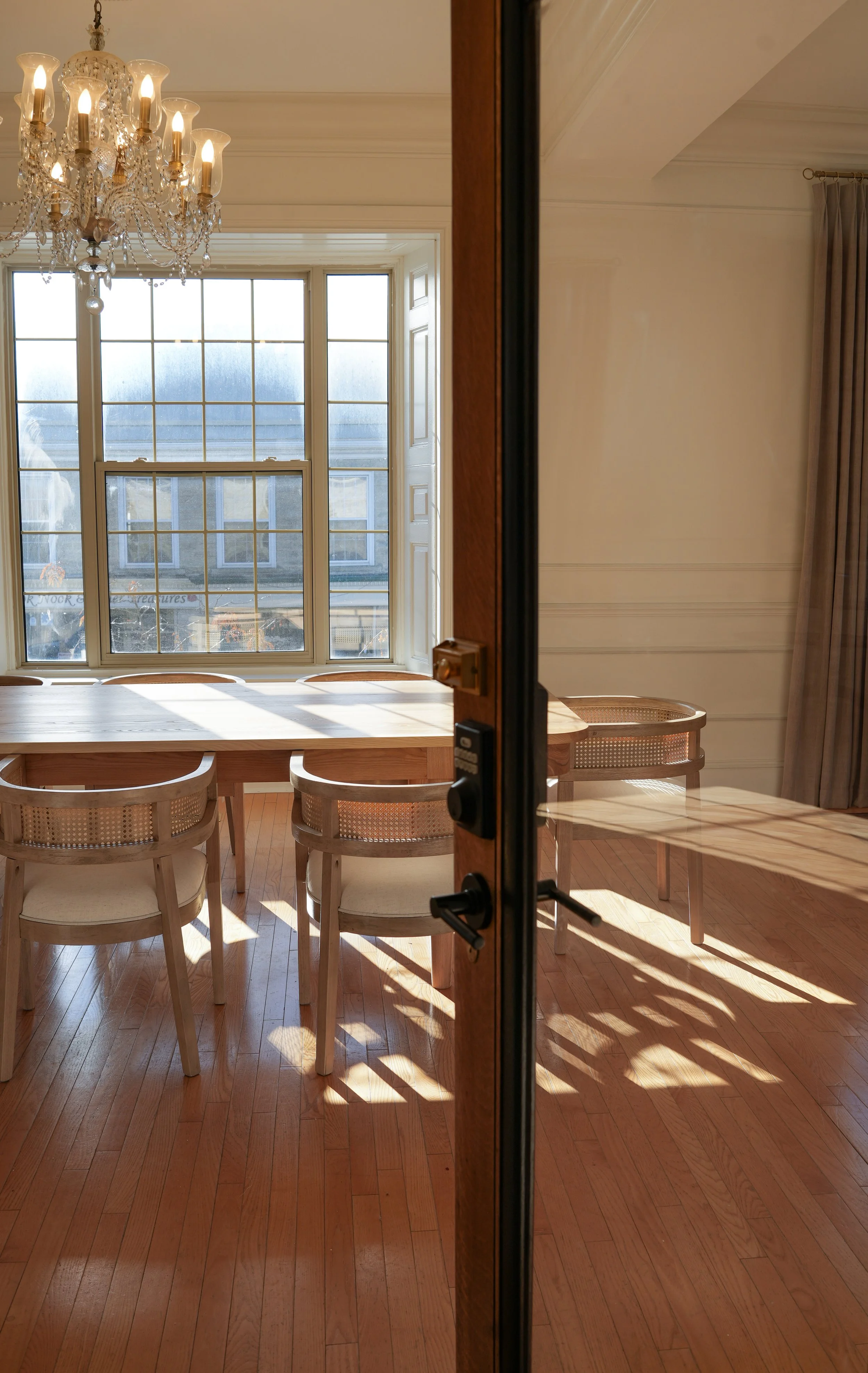 A dining room seen through a glass door, featuring a wooden table, beige chairs, a chandelier, large windows with sunlight casting shadows on the floor, and curtains on the right side.