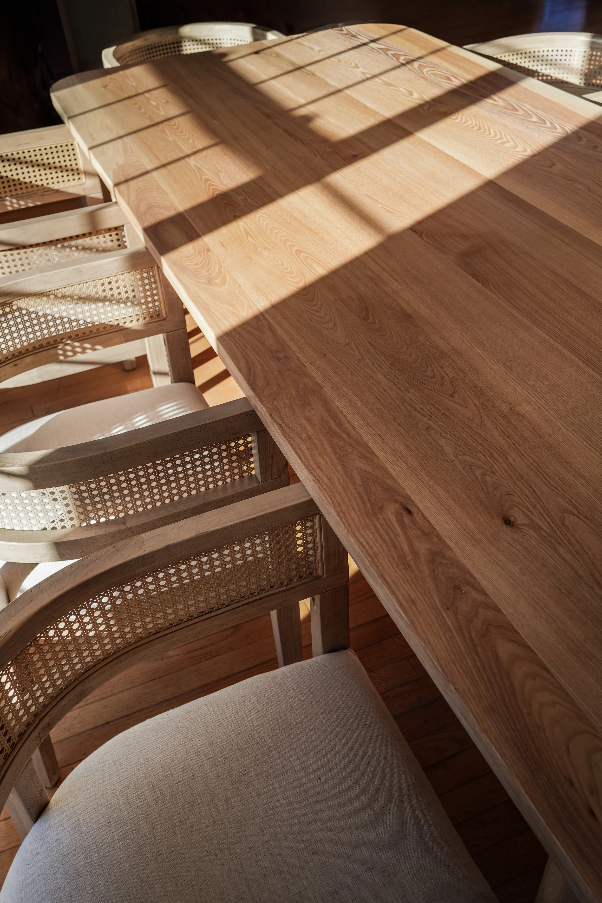 A wooden dining table with sunlight and shadow patterns, surrounded by chairs with cane backs and upholstered seats.