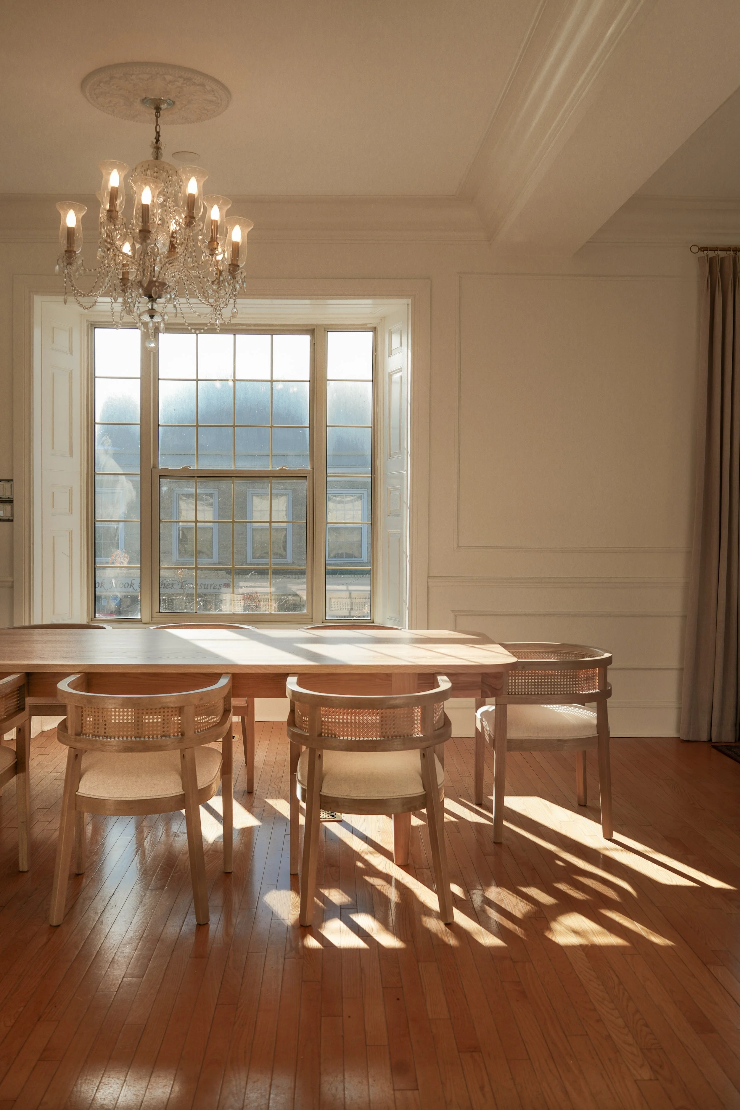 Bright dining room with a large window, chandelier, wooden table, and chairs, sunlight casting shadows on hardwood floor.