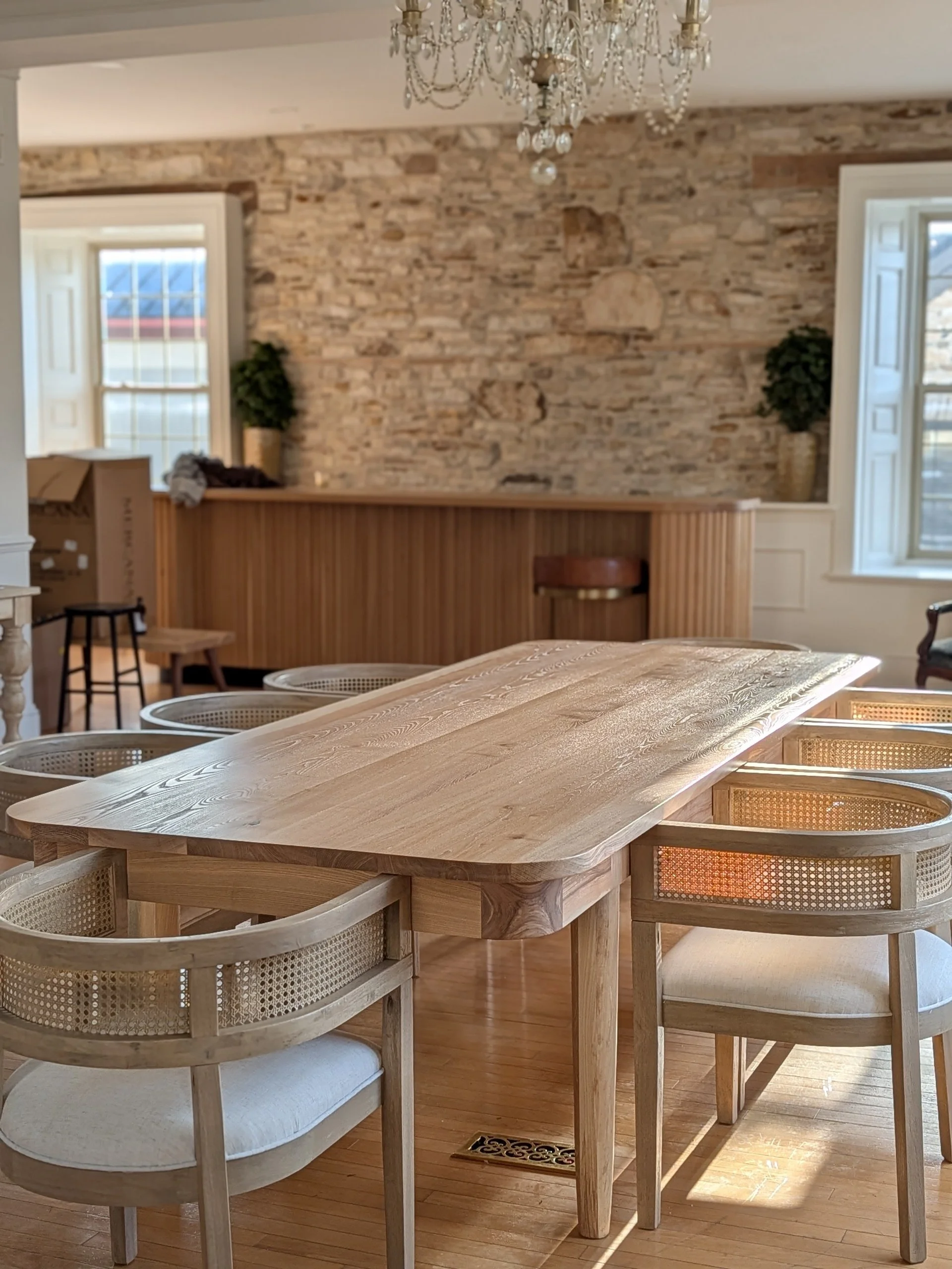 A wooden dining table with matching chairs in a room with brick walls, large windows, and a chandelier hanging from the ceiling.
