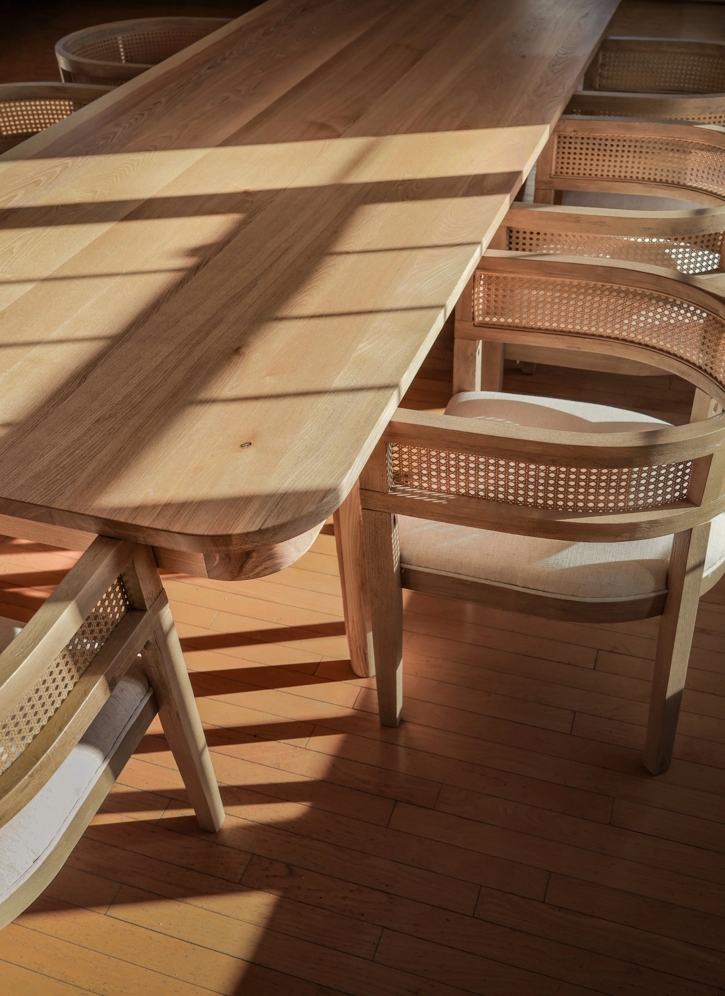Wooden dining table with four rattan-backed chairs on a hardwood floor, sunlight casting shadows.