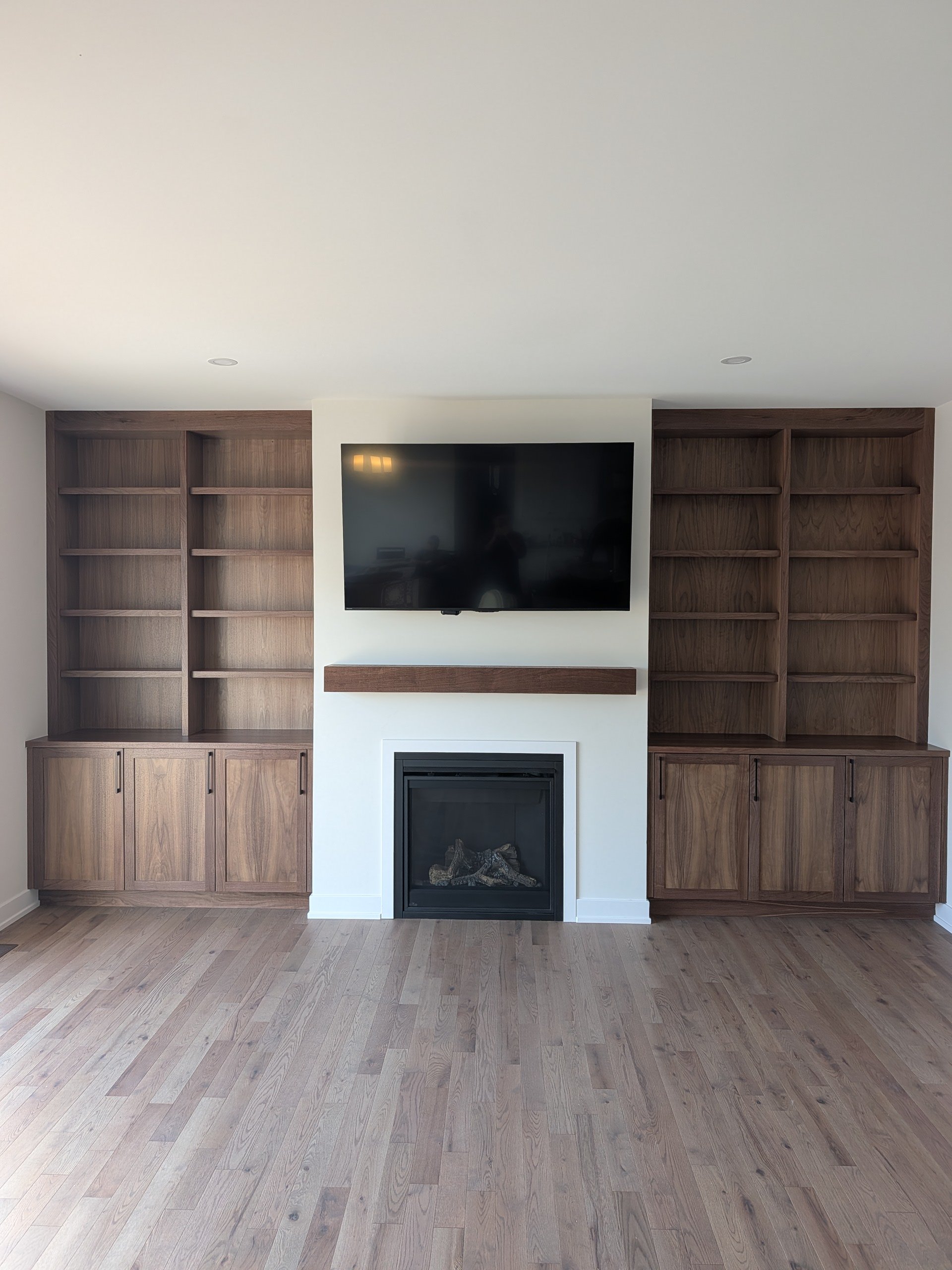 Living room with built-in wooden shelves on either side of a white fireplace, a mounted flat-screen TV above the fireplace, and wooden flooring.