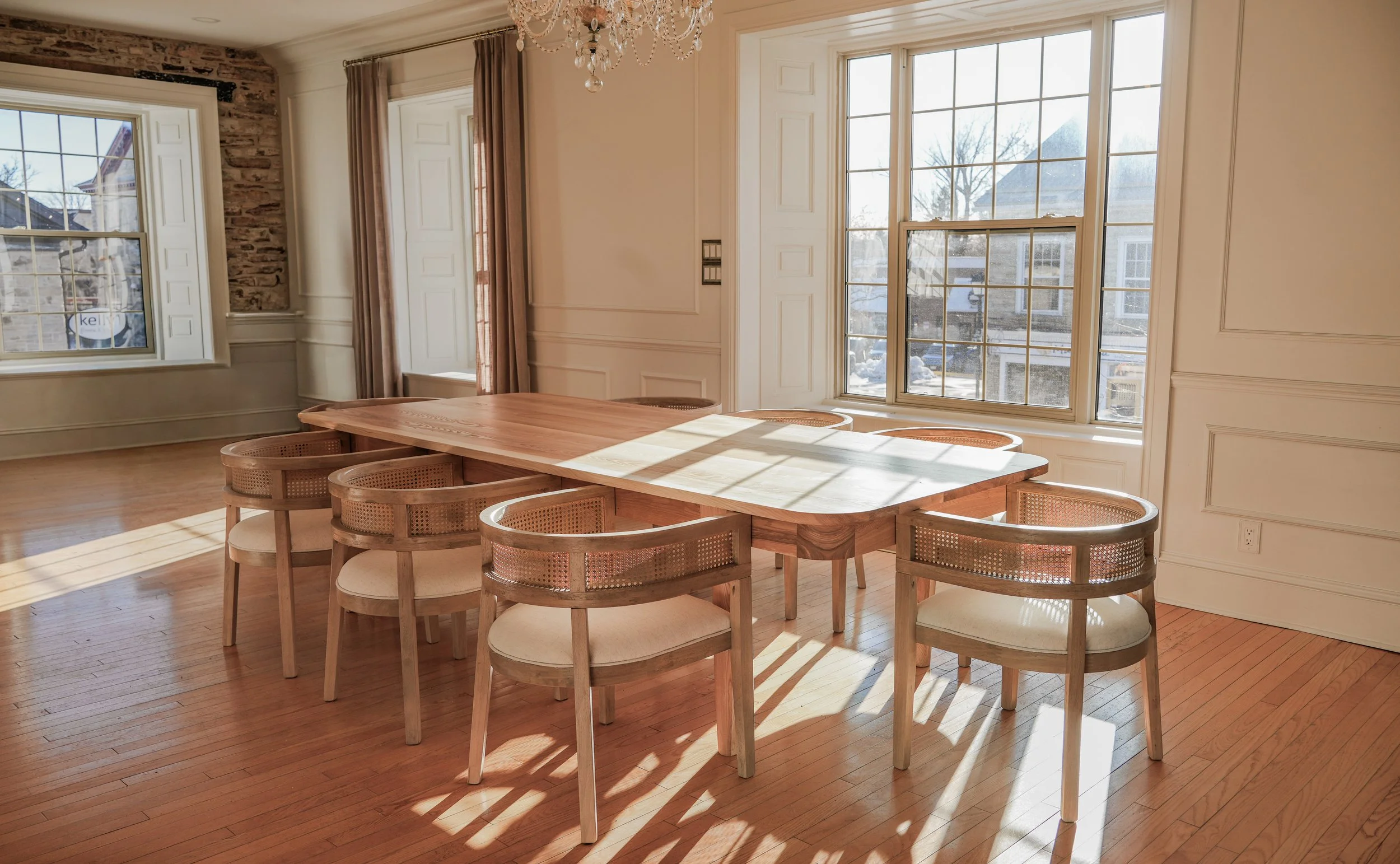 A dining room with a wooden table and six matching chairs, sunlight streaming through large windows, cream-colored walls with decorative trim, hardwood floors, a chandelier overhead, and curtains on the windows.