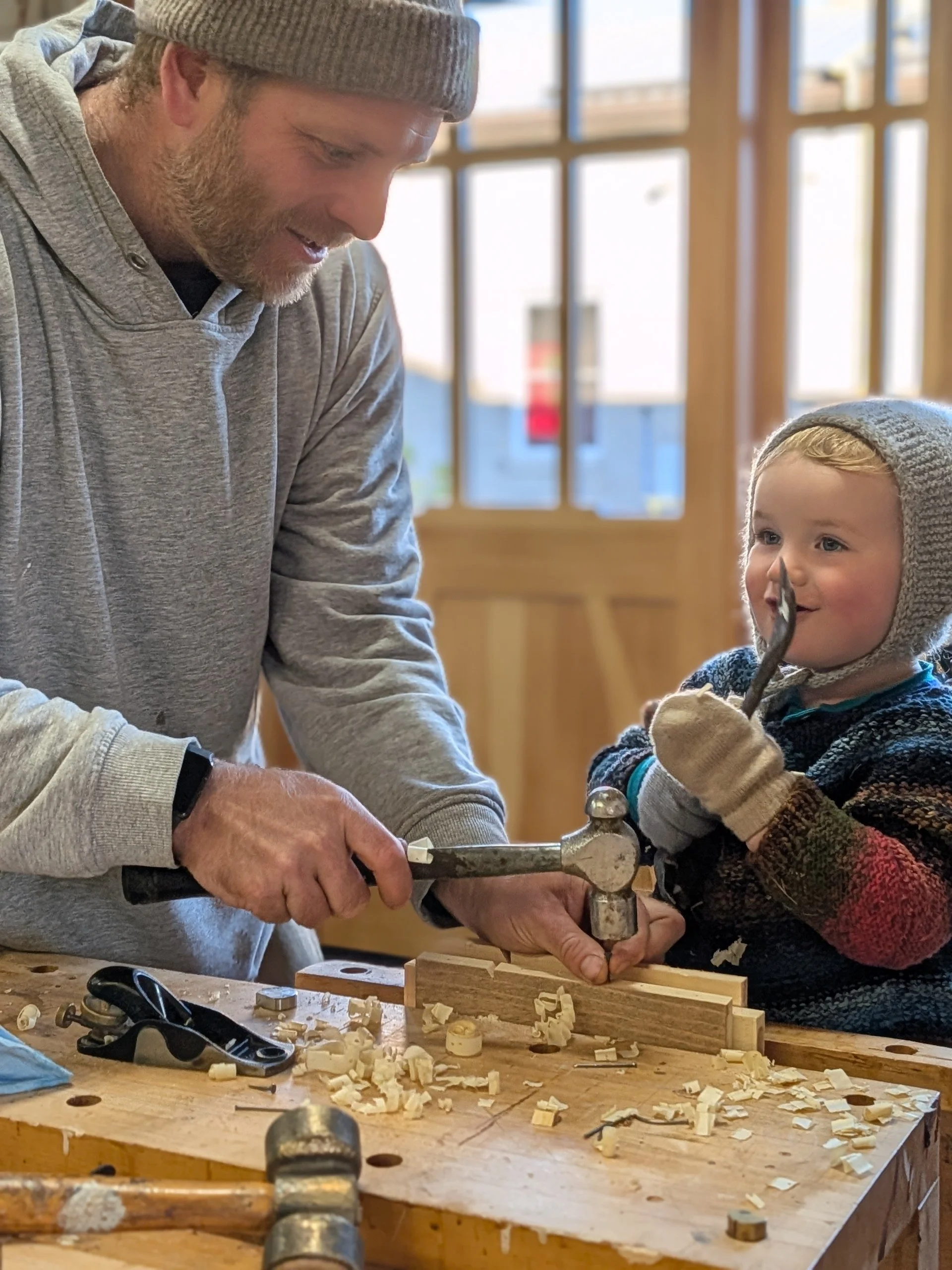 A man and a young boy working together on a woodworking project in a workshop, using a hammer and chisel on a piece of wood, with wood shavings on the workbench.
