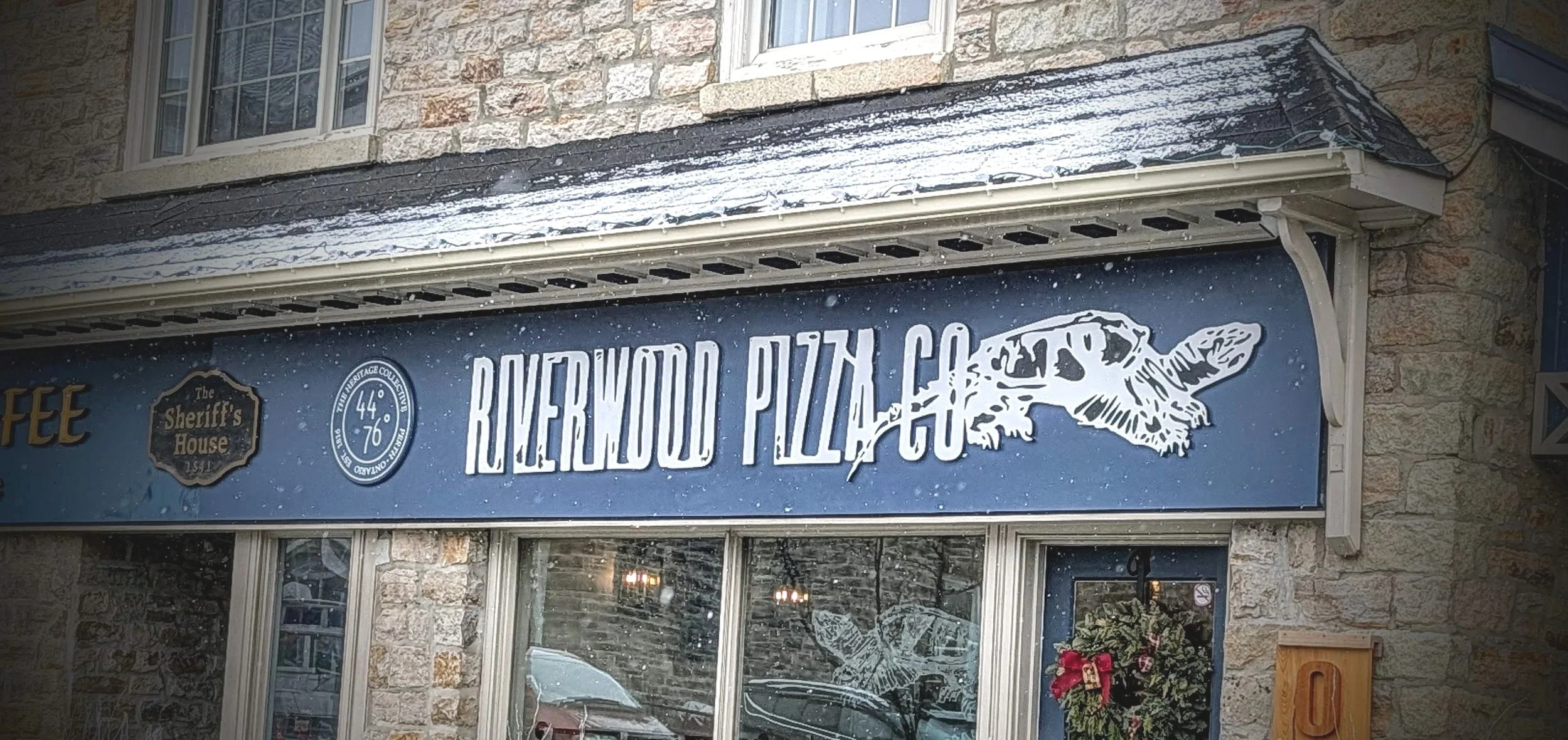 Sign for Riverside Pizza Co. with a pointing hand graphic, stars, and snow outside a stone building.