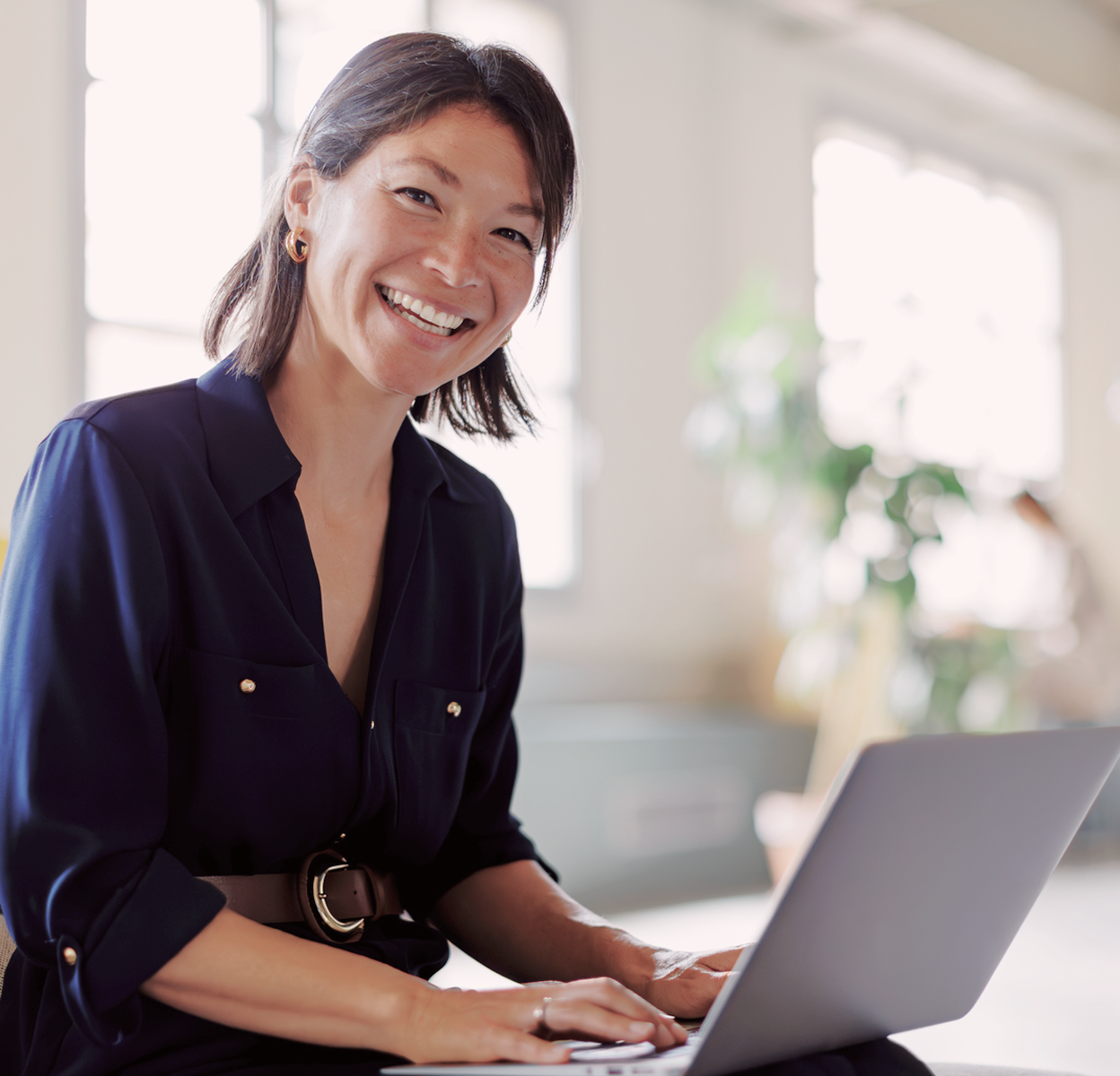 A professional smiling while working on a laptop in a bright, calm workspace.