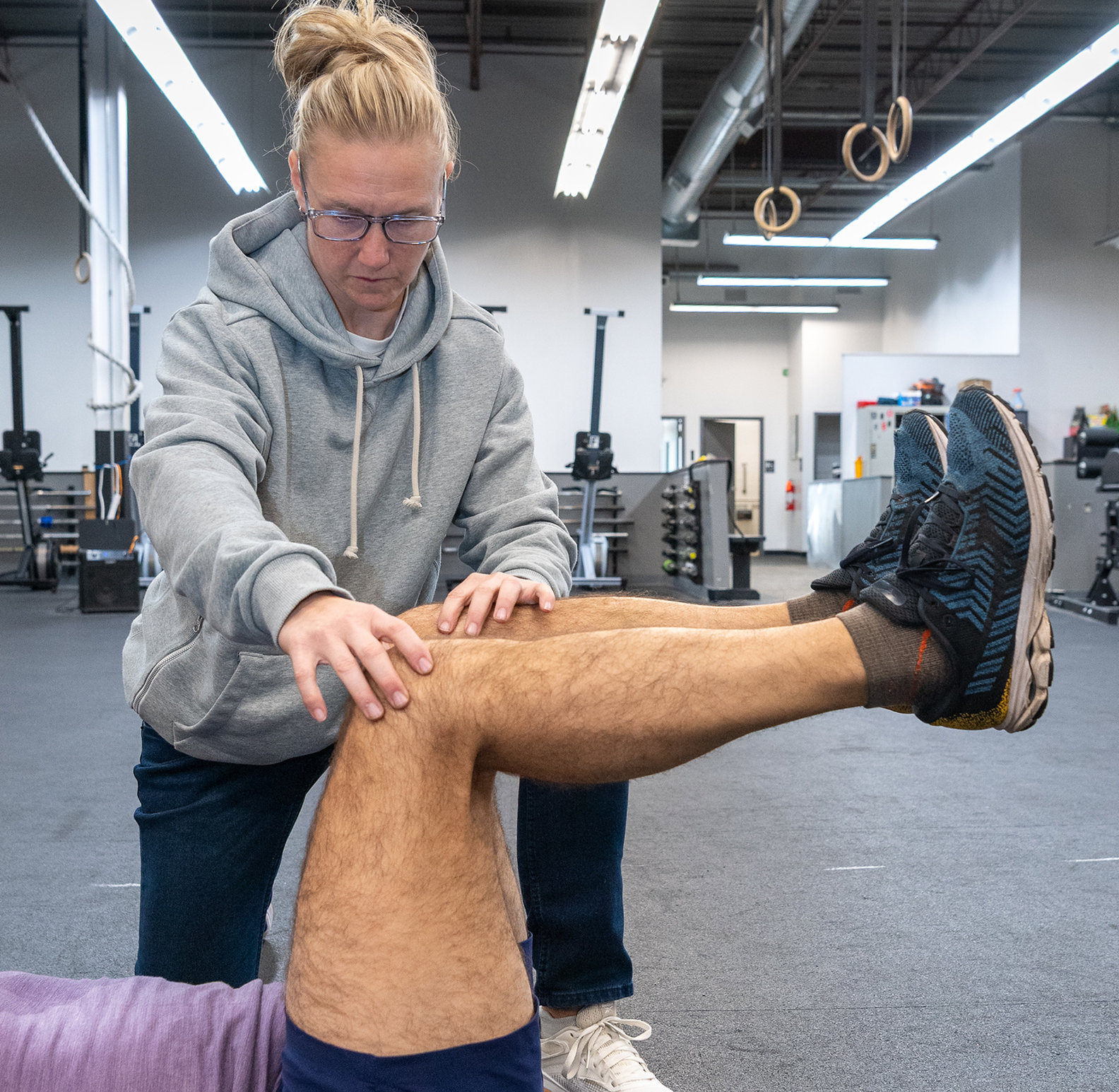 A professional assessing a person’s leg position during a guided movement exercise in a gym setting.