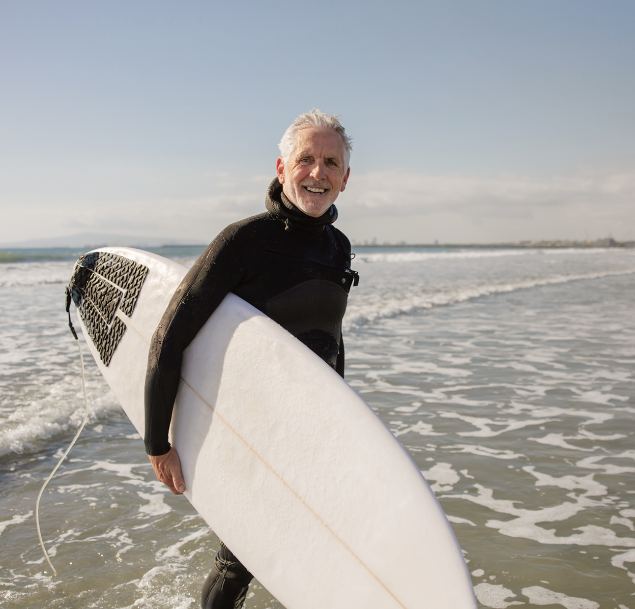 An older adult carrying a surfboard at the shoreline, smiling and active in the ocean.