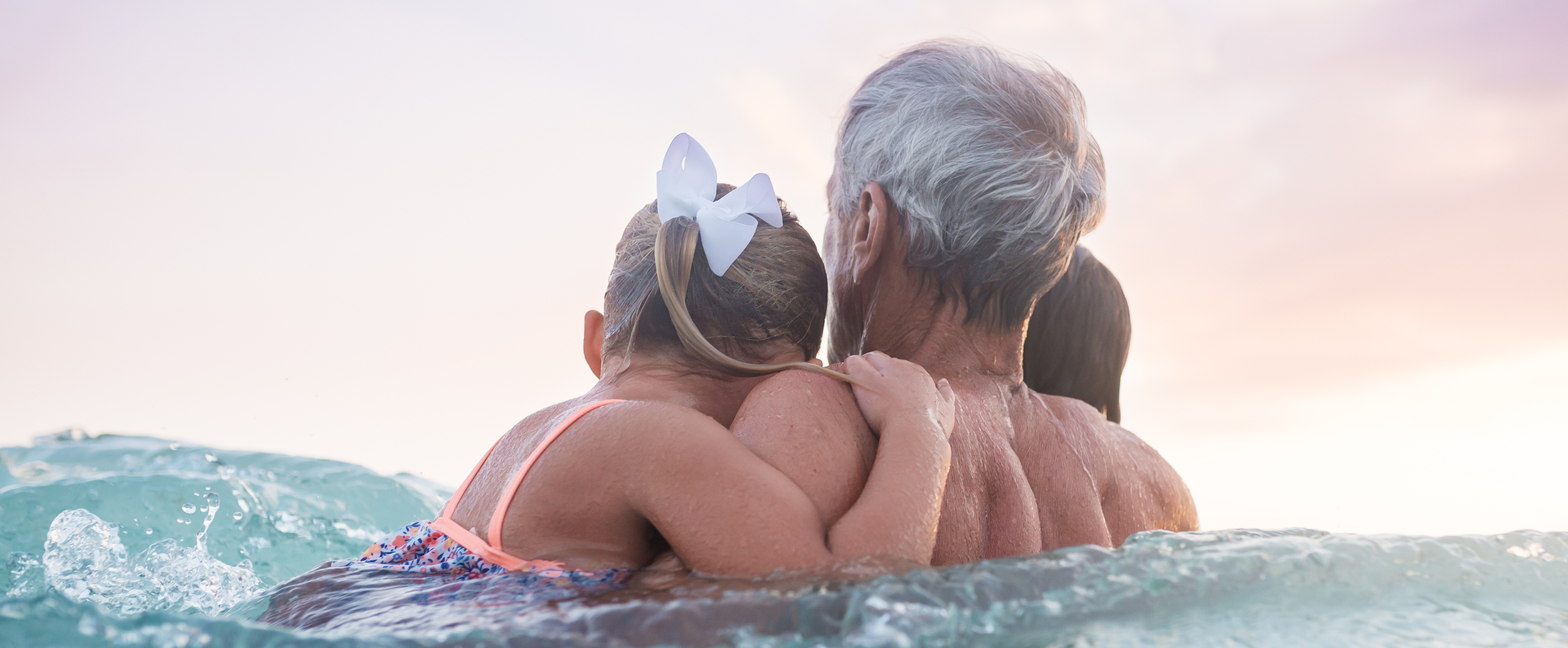 An older adult holding a child in the ocean, seen from behind, sharing a calm, supportive moment in the water.