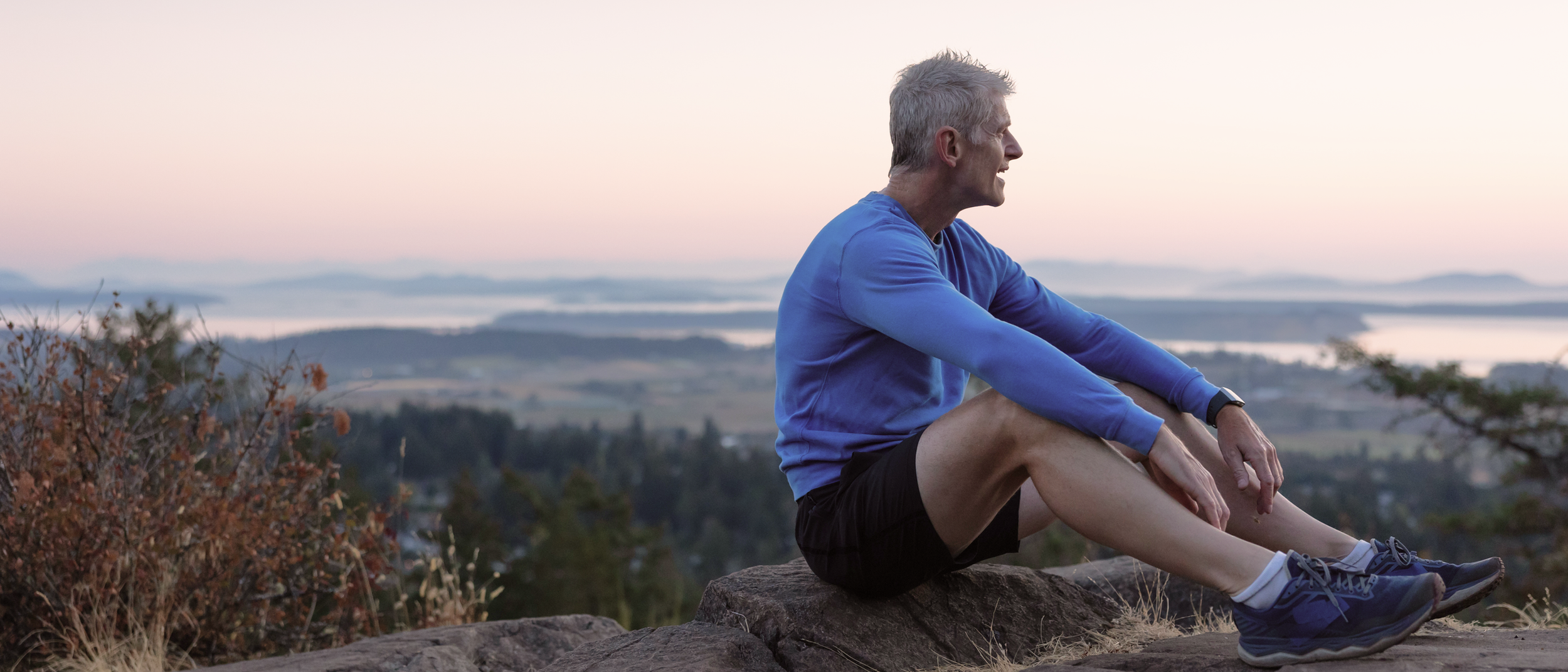 An older adult sitting on a rock at sunset, resting after exercise and looking out over a wide landscape.