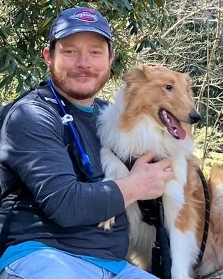 A man wearing a baseball cap and a long-sleeve shirt sitting outdoors with a fluffy Shetland Sheepdog.