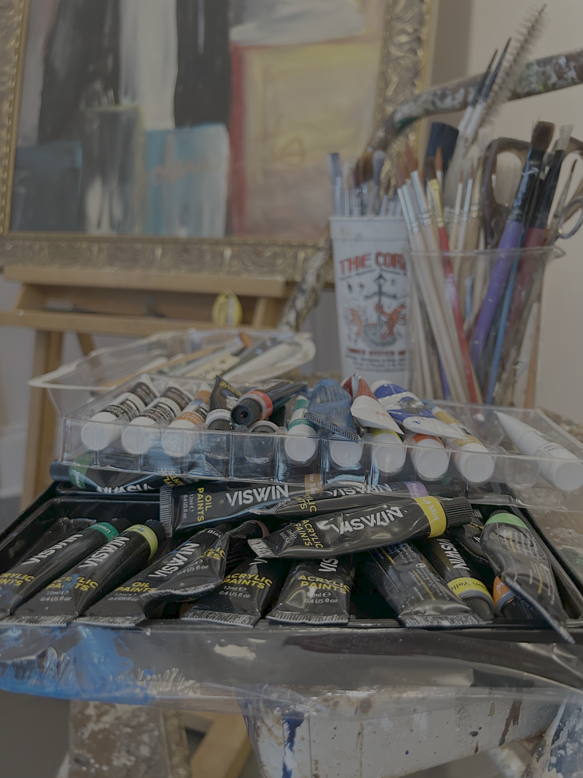 A box of acrylic paint tubes and a container holding paintbrushes on table at john malta's studio with an easel and a painting in the background.