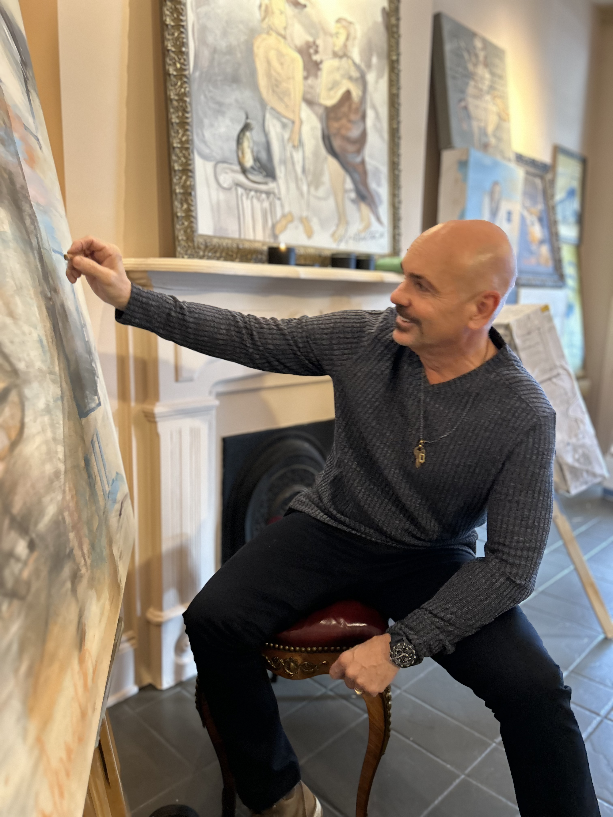 A man with a bald head and a goatee, wearing a dark gray long-sleeve shirt, is sitting on a red upholstered chair and pointing at a painting on an easel. The scene appears to be an art gallery or studio with multiple paintings displayed on the walls in the background.