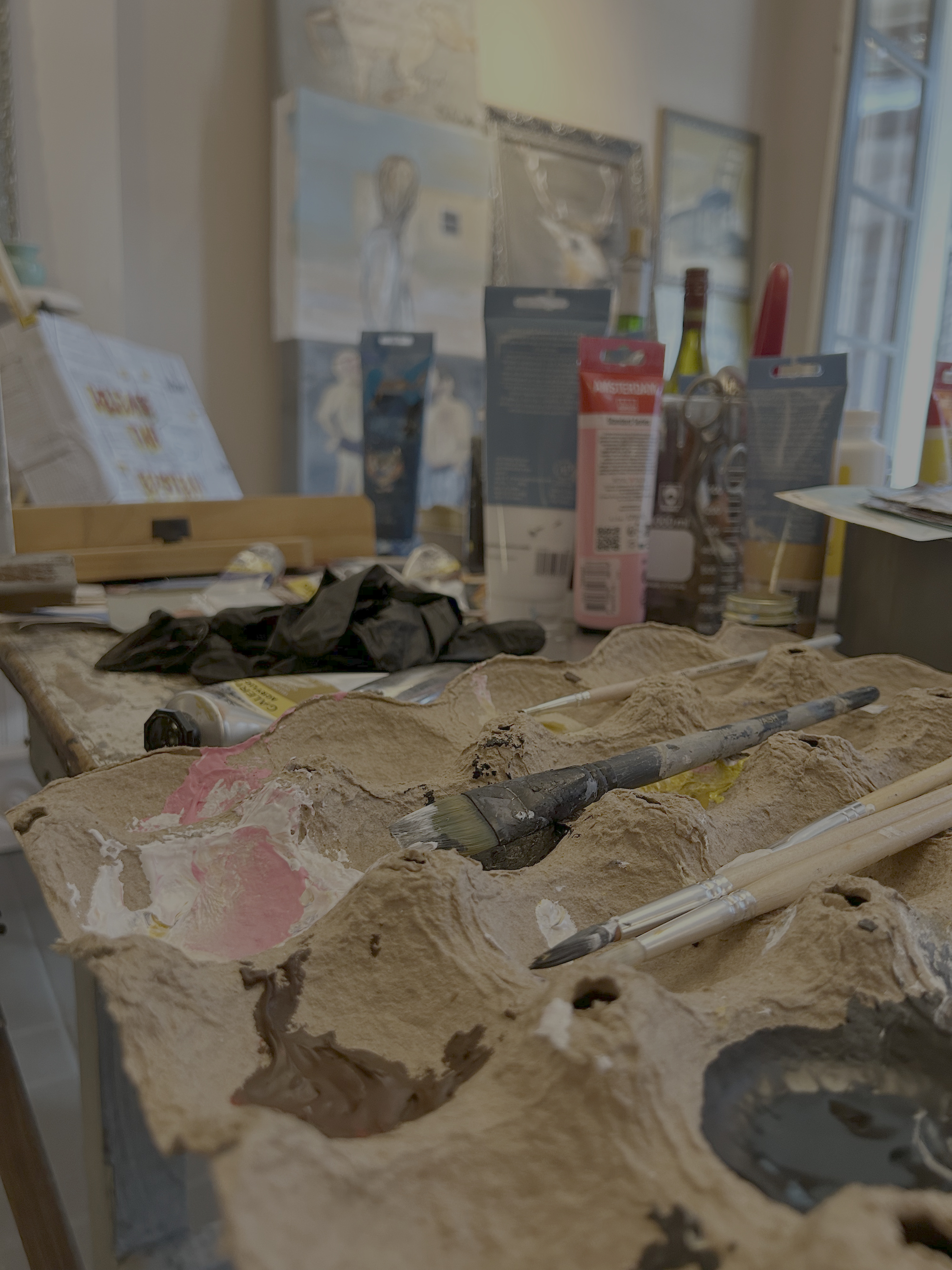 john malta's clay palette with paint and brushes sits on a table, with art supplies and paintings visible in the background.