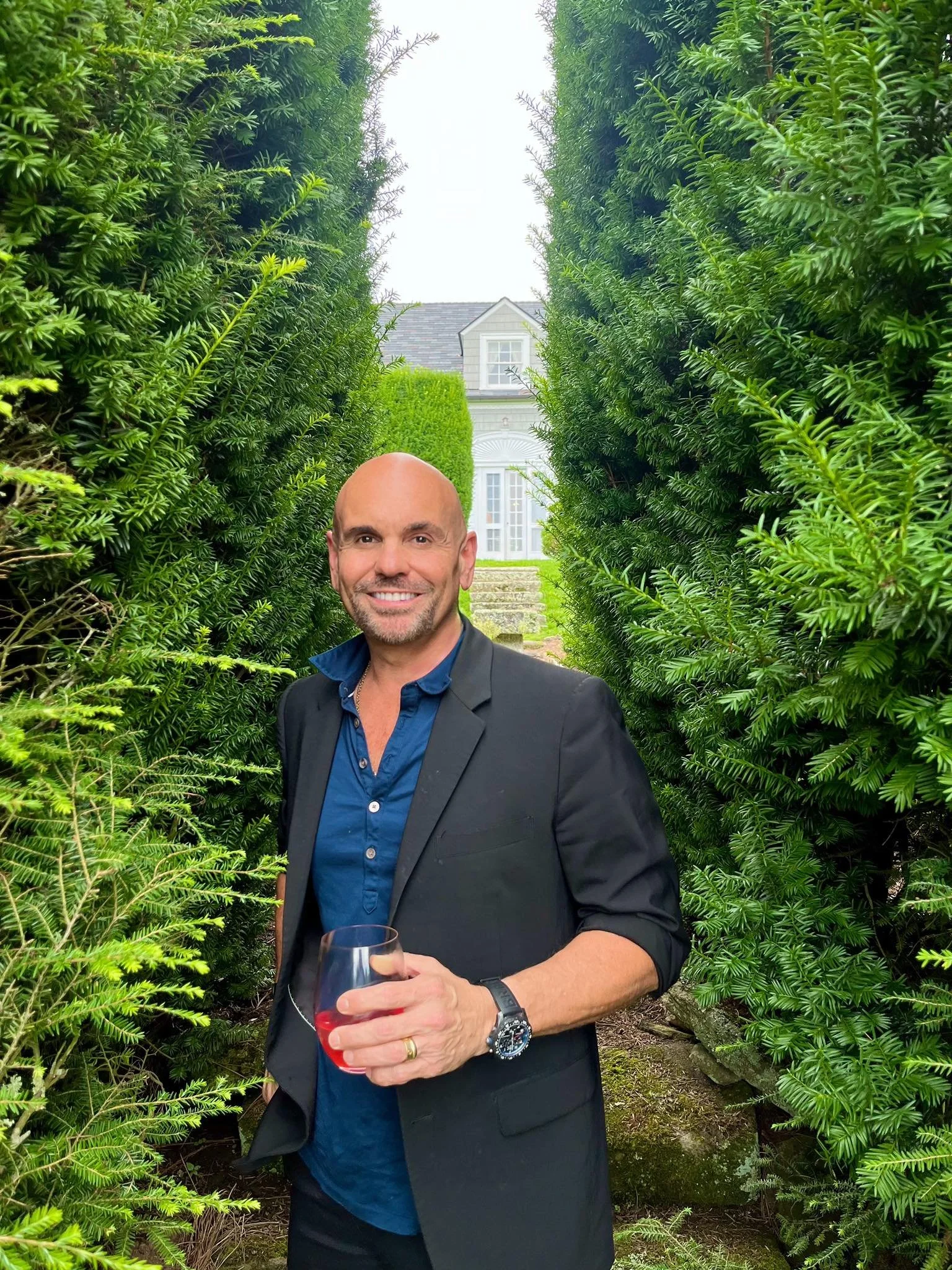 john malta holding a glass standing between tall green bushes, with a white house visible in the background.