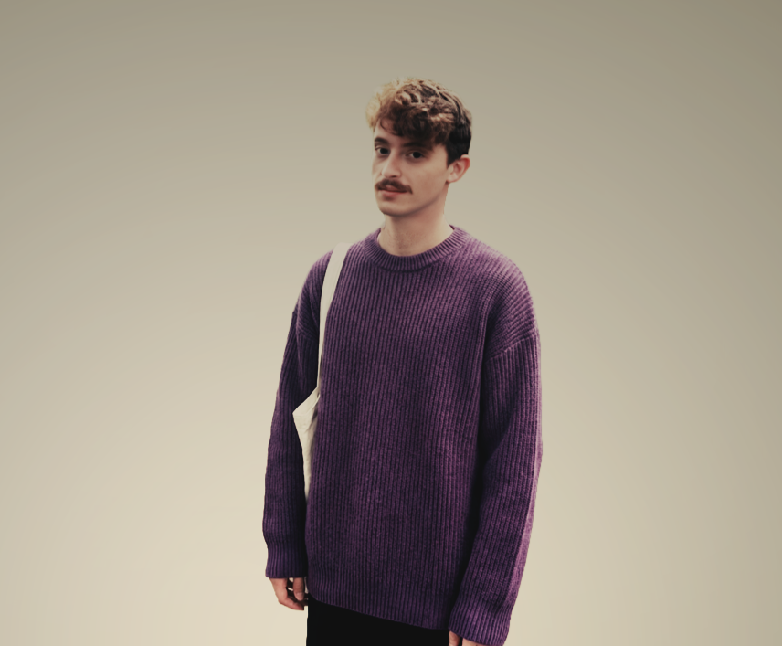 A young man with curly brown hair and a mustache stands against a plain, light-colored background. He is wearing a purple knit sweater and carries a white shoulder bag.