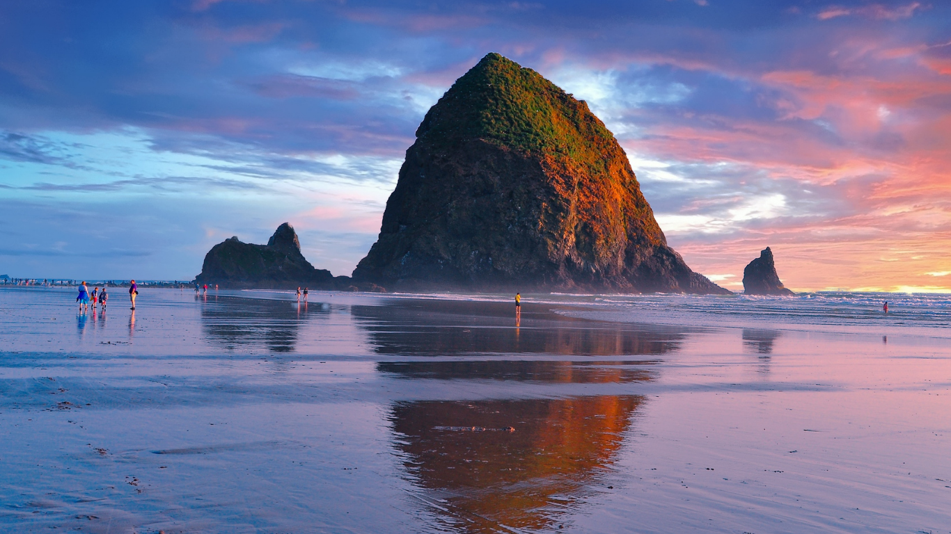 People walking on the wet sandy beach with Haystack Rock and smaller rock formations in the background during sunset.