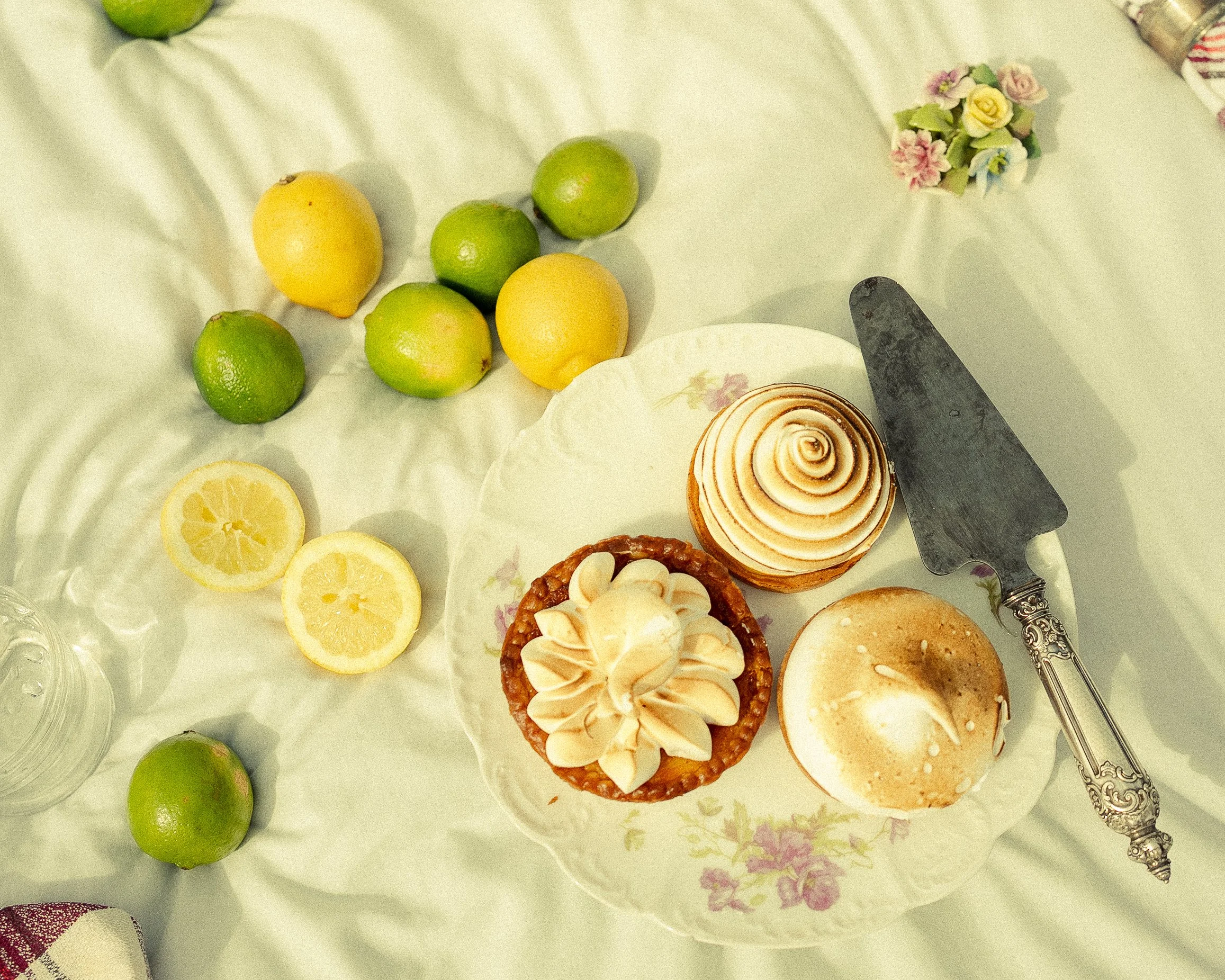 A table set with a plate of two lemon cupcakes, a lemon tart, and a meringue-topped dessert, along with whole and sliced lemons and various limes on a light-colored tablecloth.