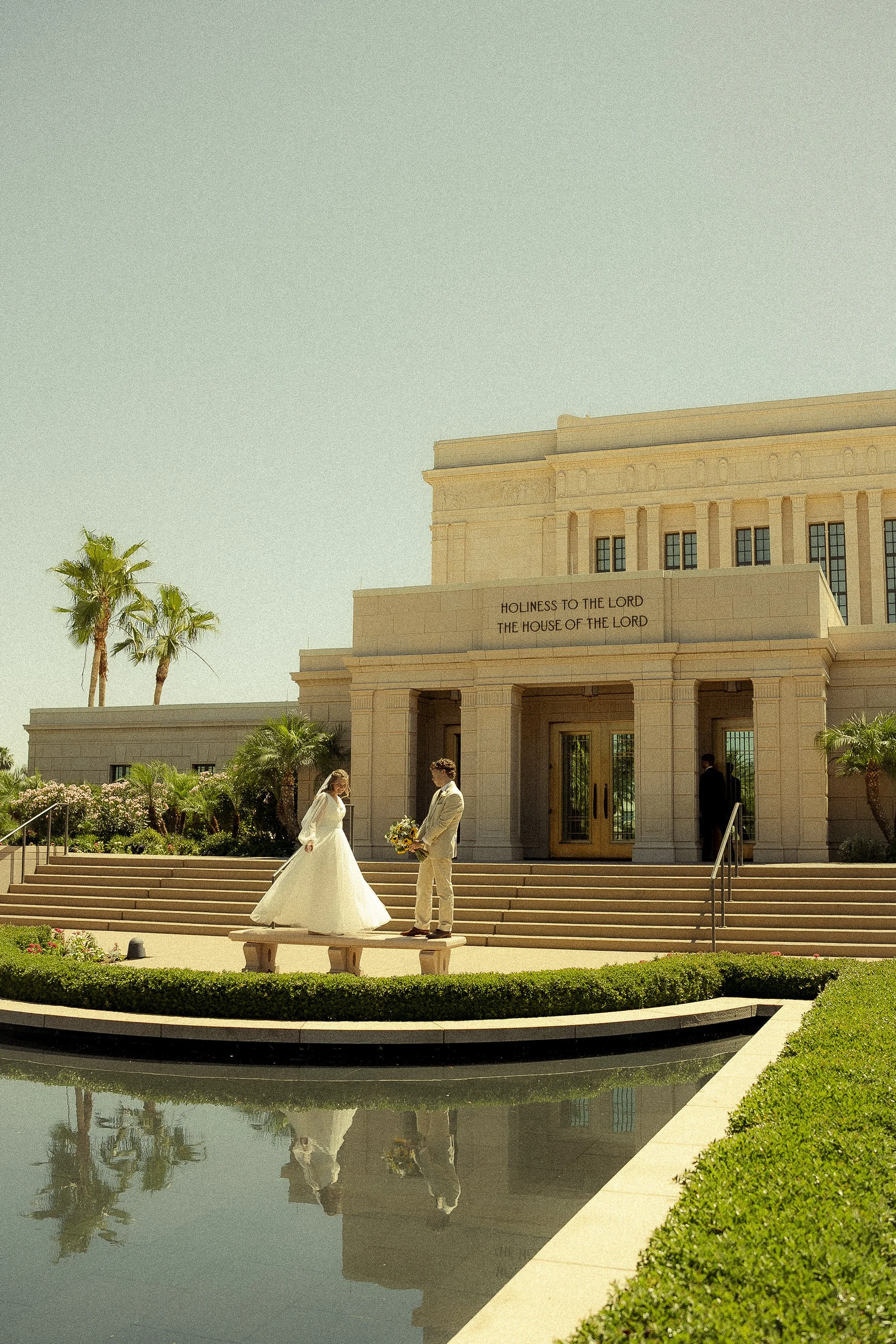 Un couple habillé en vêtements de mariage se tient devant un bâtiment imposant avec une inscription en anglais. La scène se déroule devant une fontaine avec un reflet dans l'eau, entourée de verdure et de palmiers.